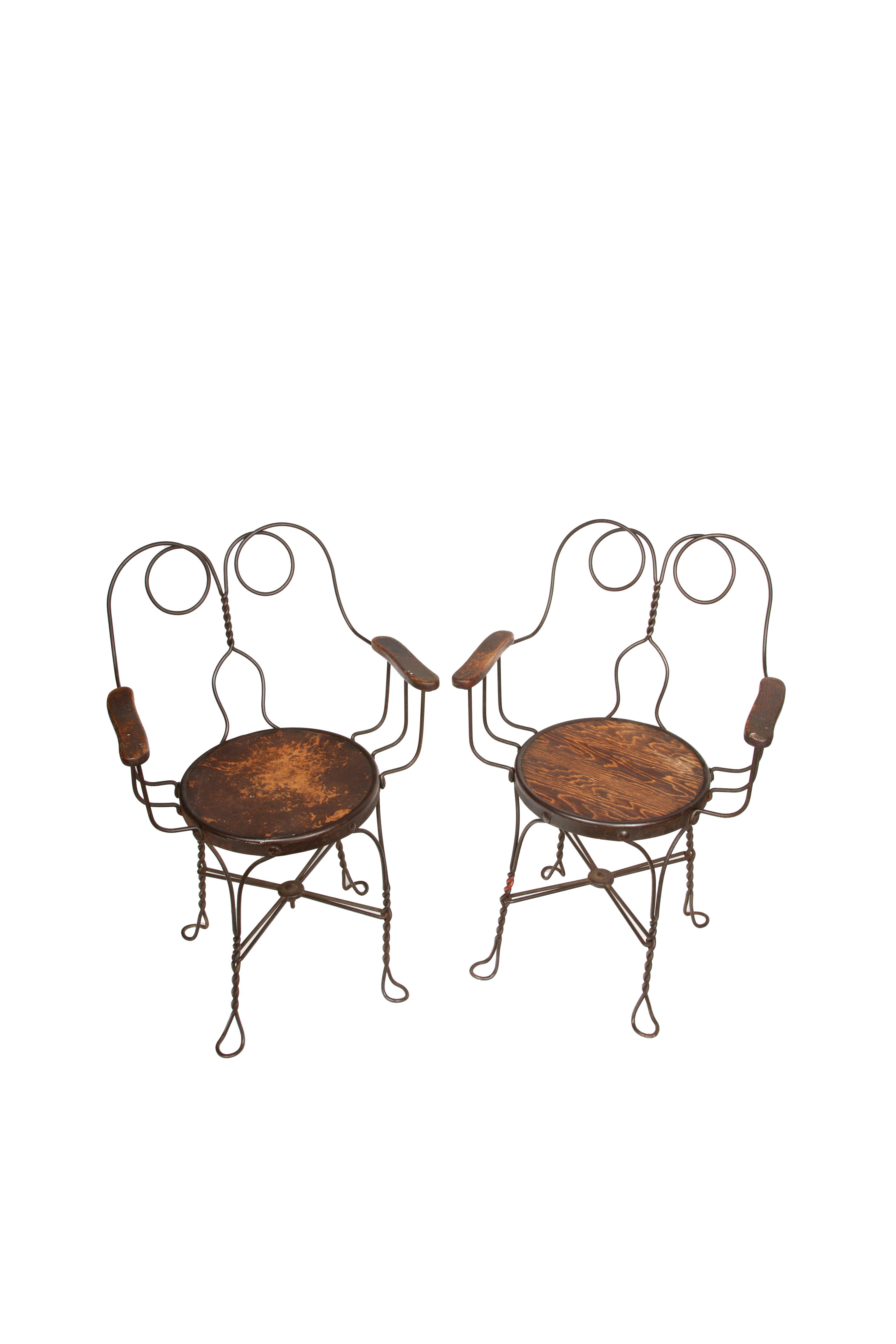 This charming pair of antique ice cream parlor armchairs, dating from the early 1900s, features an elegant twisted wrought iron frame that is both sturdy and visually appealing. The chairs are characterized by their distinctive scrolled backrests