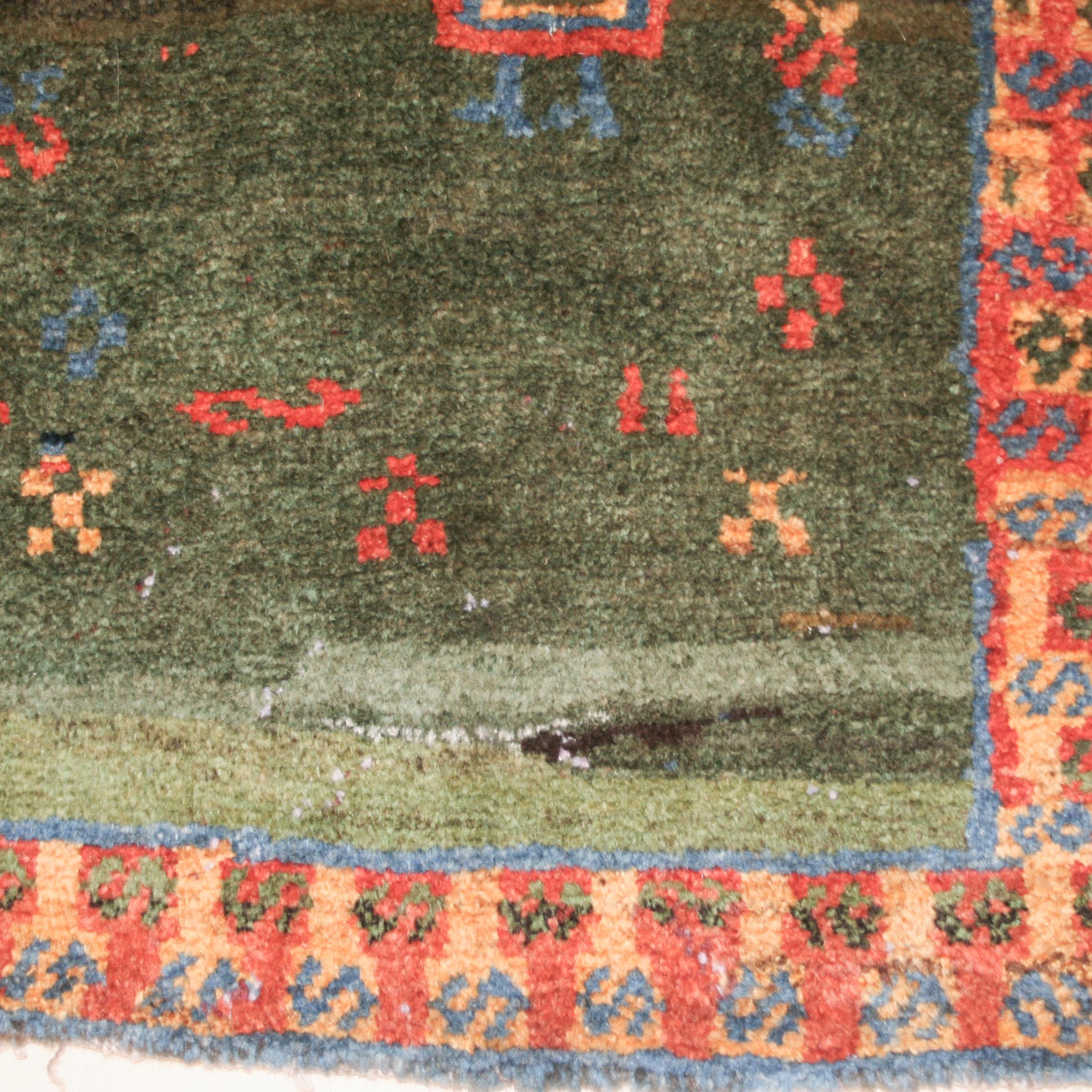 Un tapis tribal très inhabituel, qui se distingue par un fond vert émeraude en divers dégradés, agrémenté de motifs géométriques épars ainsi que d'une figure anthropomorphe. La bordure jaune du fond contient un ensemble parallèle d'éléments en forme