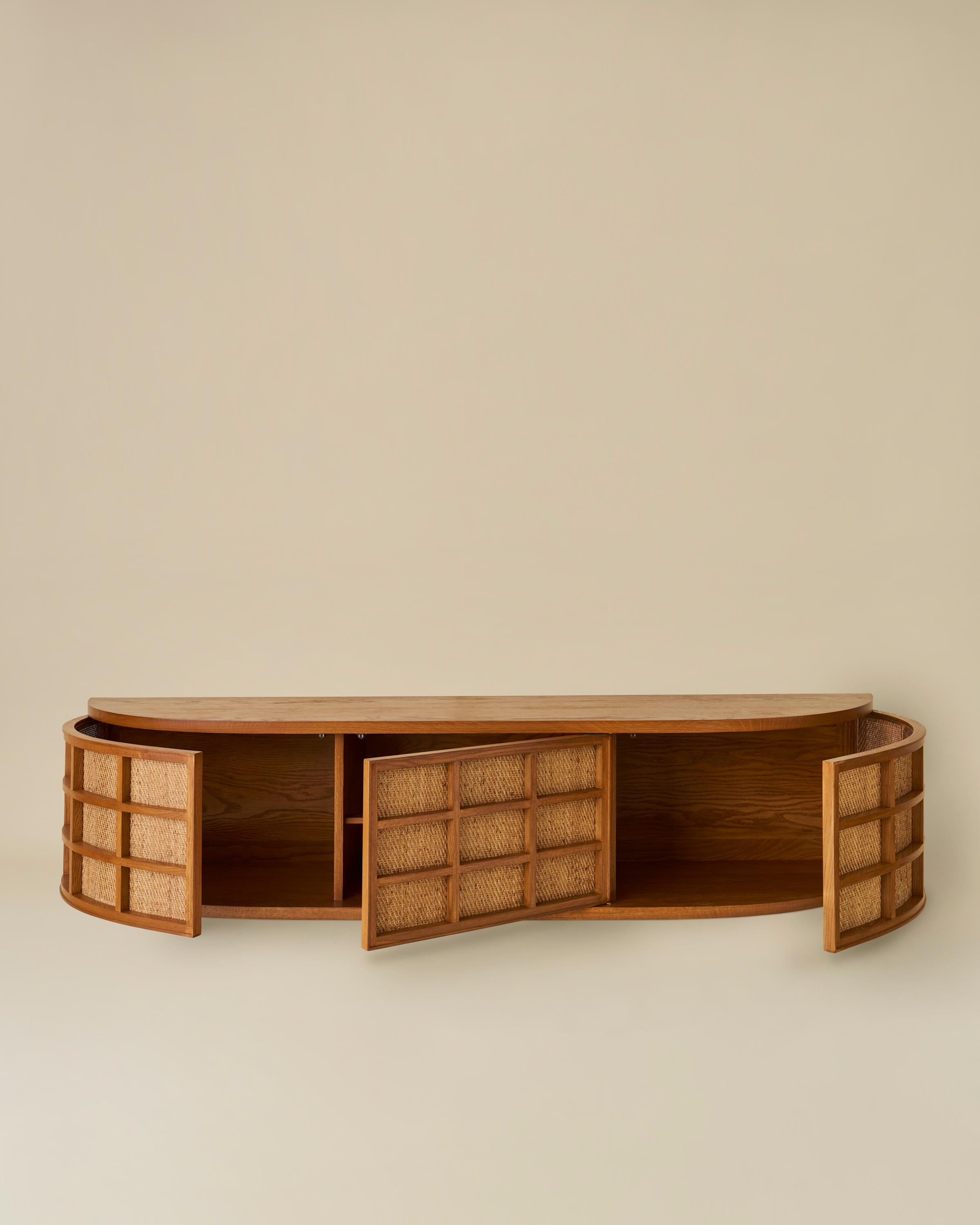 Une grille formée en chêne massif enveloppe la façade de cette crédence murale flottante.  Trois portes à fermeture par pression révèlent des espaces de rangement et une étagère centrale.  Représenté en chêne miel avec panneaux de cannage naturel.