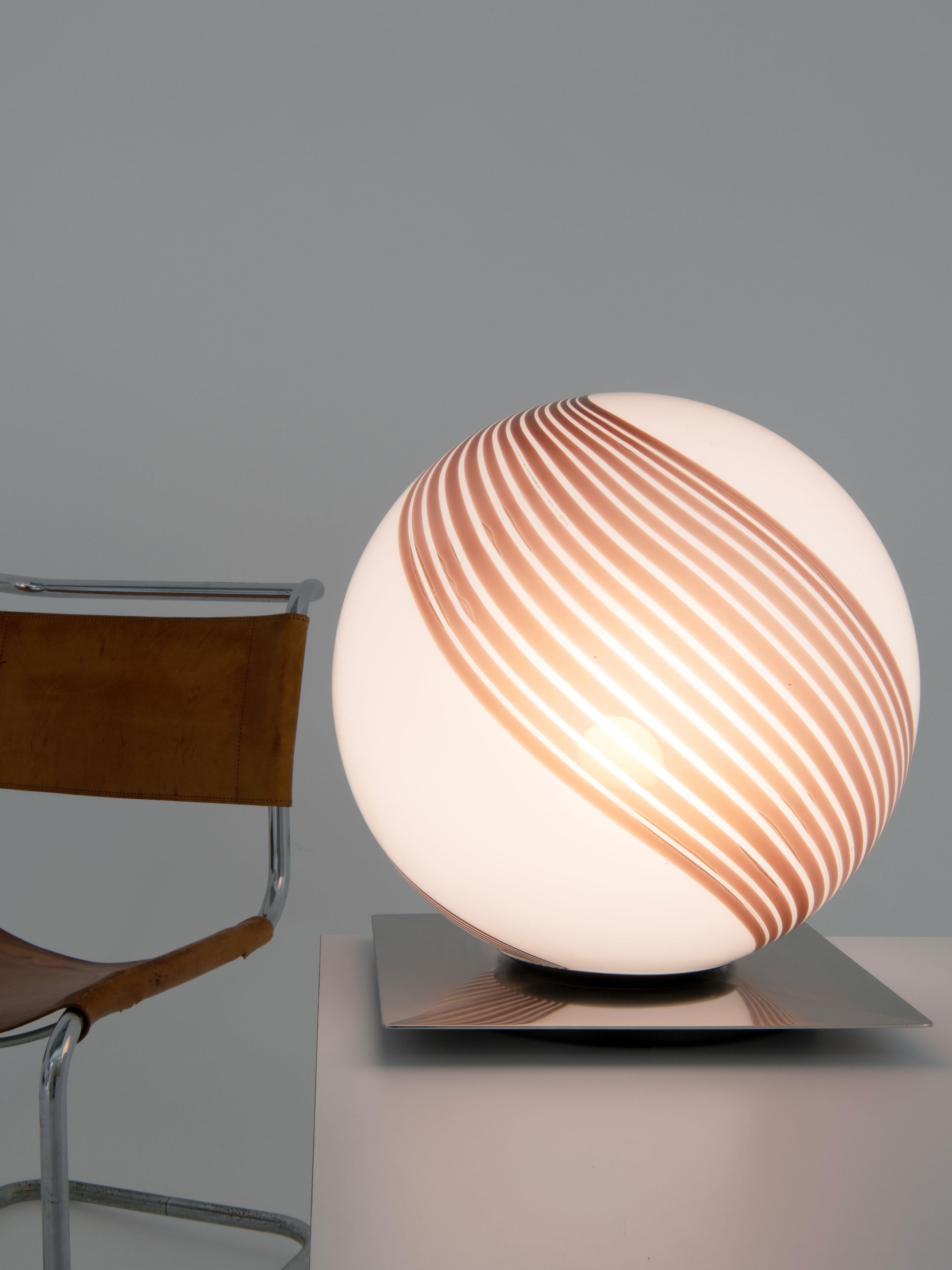 This table lamp was designed and manufactured by Lamperti, an Italian company specialising in lighting.
The lamp is made of Murano glass and rests on a sloping square base of brushed stainless steel. The swirl pattern differs from other existing