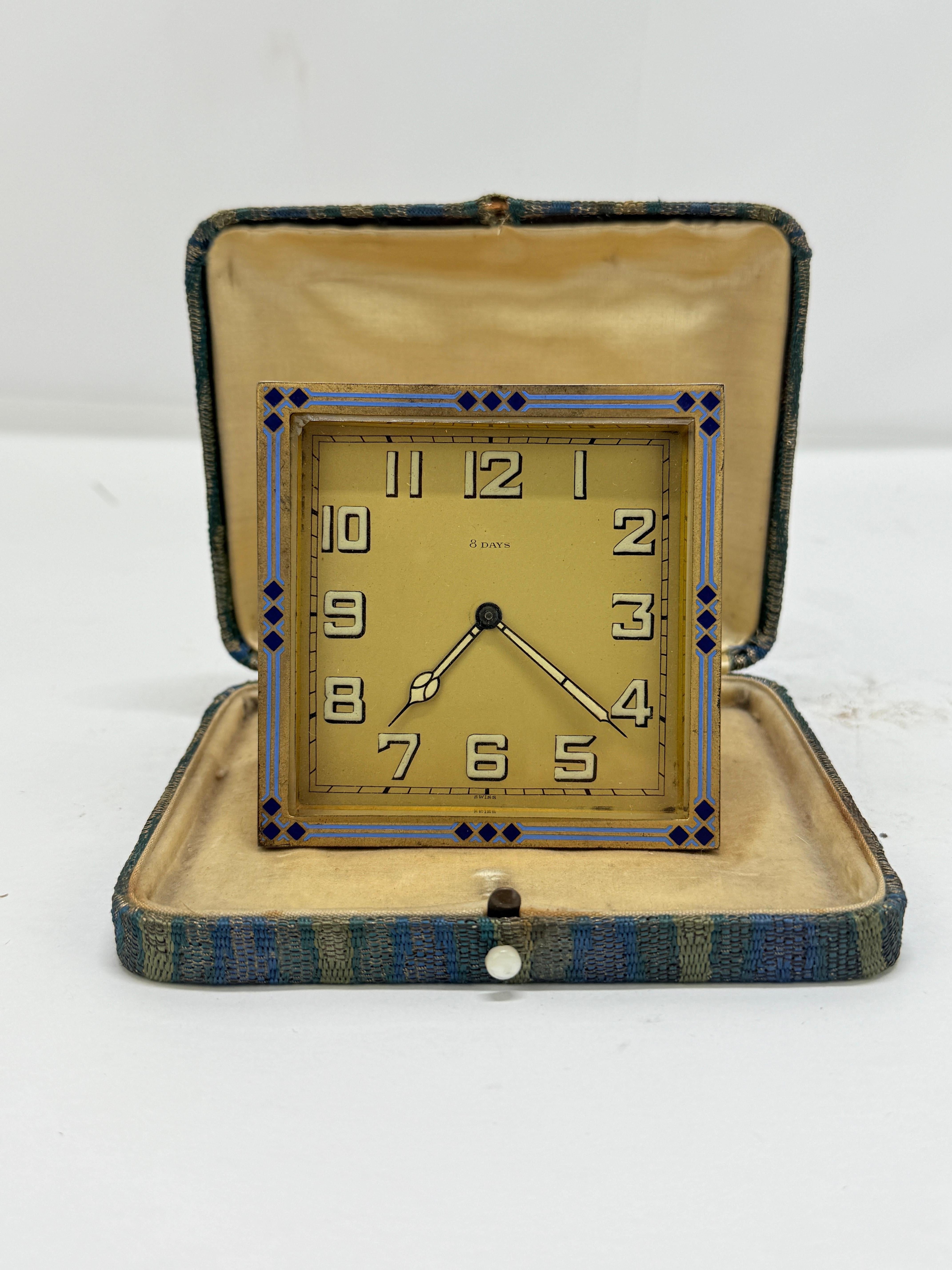A refined small-format Art Deco desk clock with an unmistakably Swiss 8-day movement, presented in a square gilt-bronze case with a fold-out strut back. The front is framed by a crisp geometric border of kiln-fired enamel in two tones of