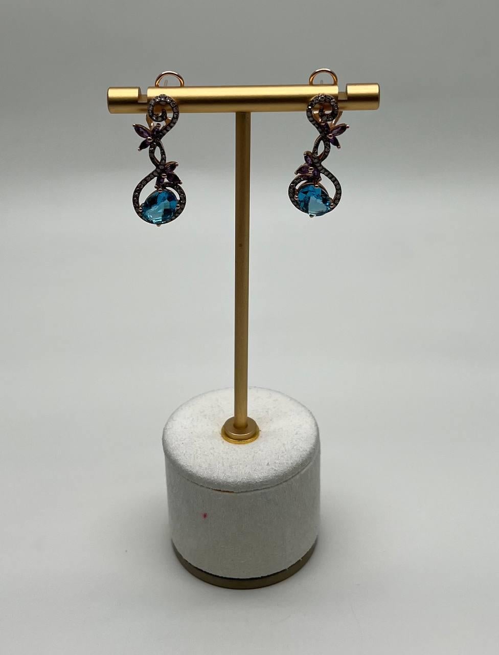 These exquisite earrings feature a delicate intertwining design adorned with vibrant blue topazes and purple accents. The intricate loops are encrusted with shimmering stones, adding a touch of sparkle. The earrings are crafted with precision,