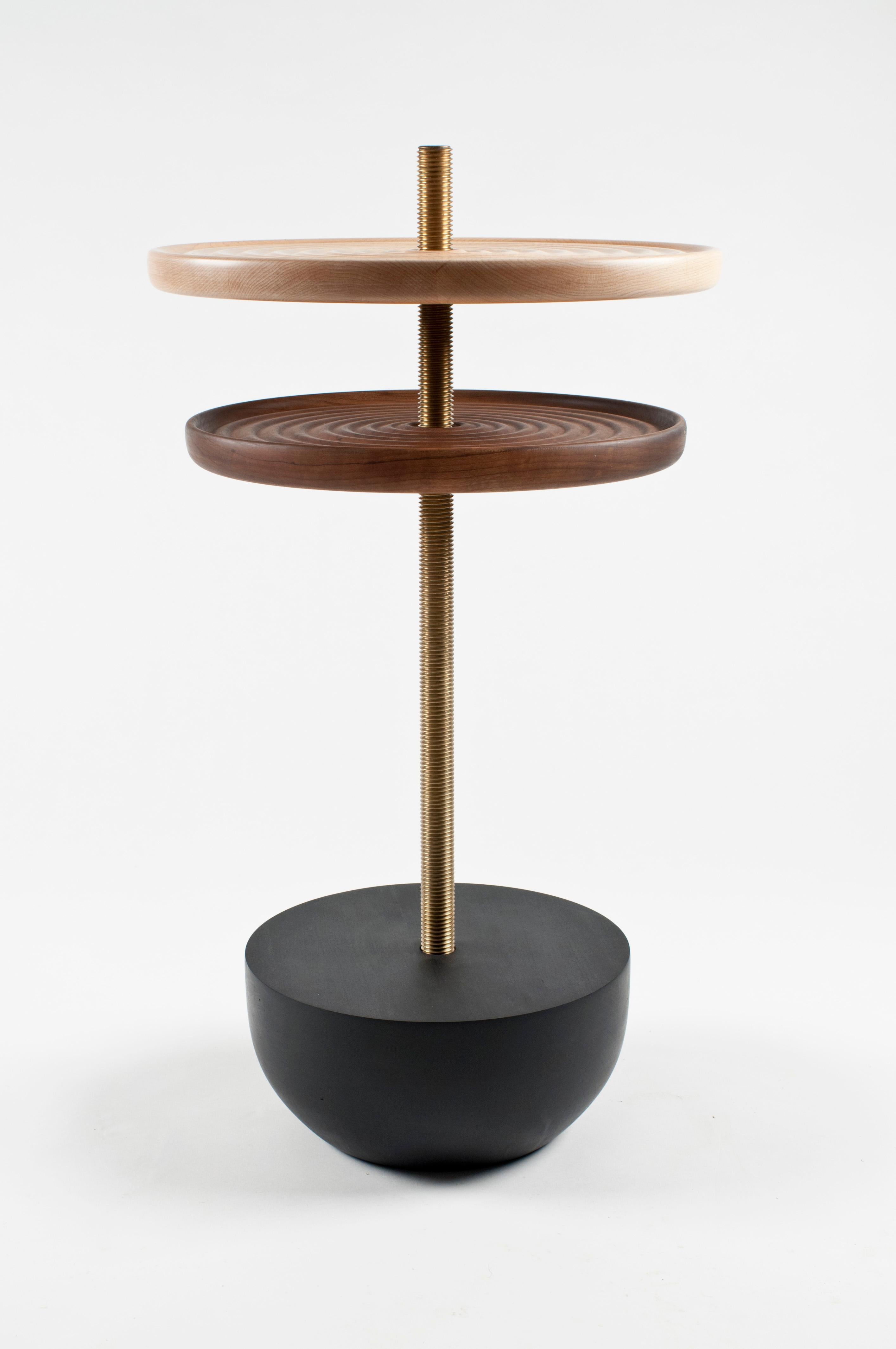 The Swivel table is handcrafted in cherry, black walnut, and brass. Each hand turned solid wood top can independently spin up and down the solid brass threaded column. Each top is double sided; the ripple textured side is visually striking and