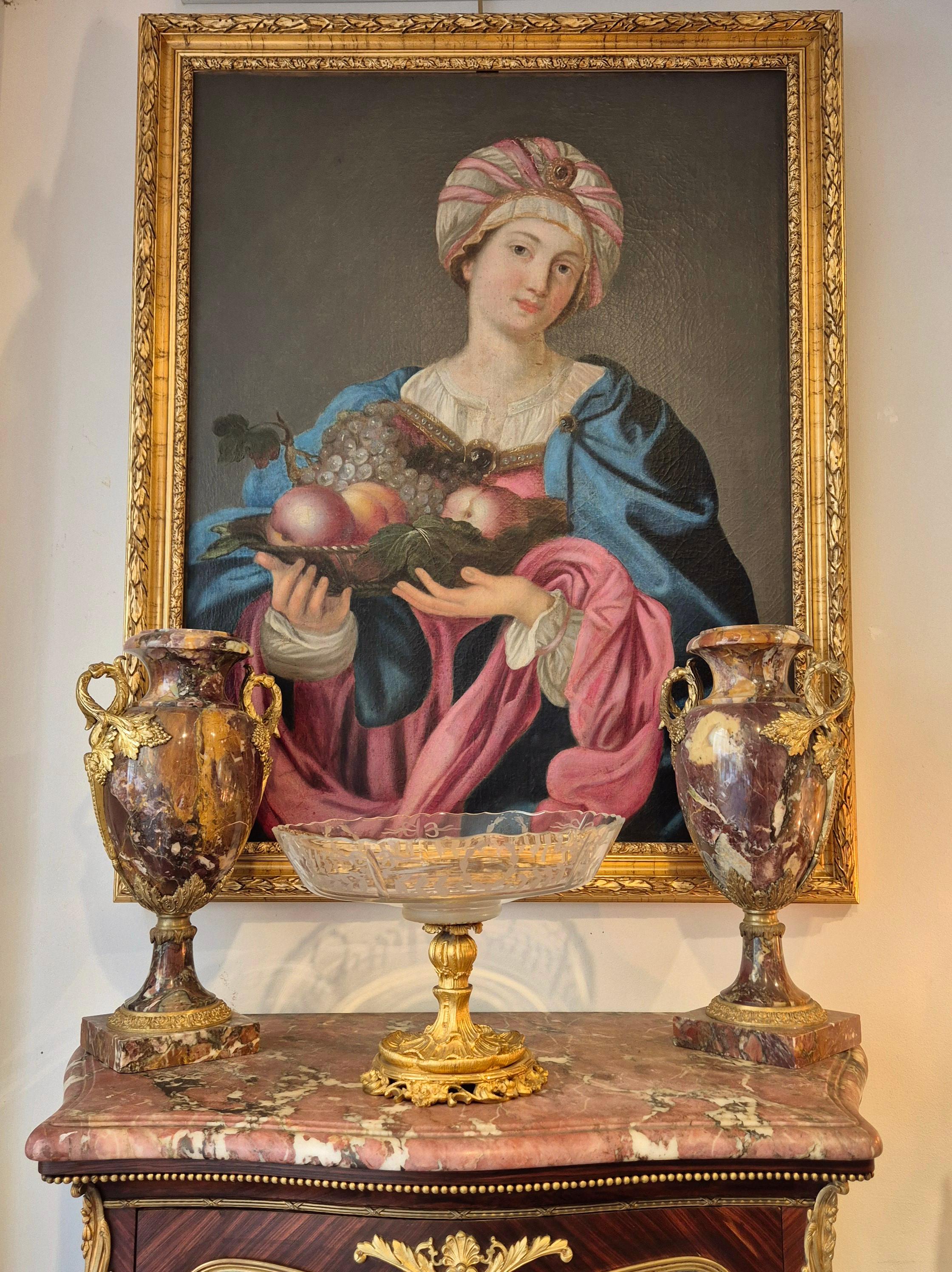 This sensual painting depicts a silk-turbaned Sybille carrying a bowl of fruit (peaches and grapes) and richly dressed in a light white linen chemise, a luxurious fuchsia-pink tunic and a royal-blue cape. She is adorned with gold jewels and precious