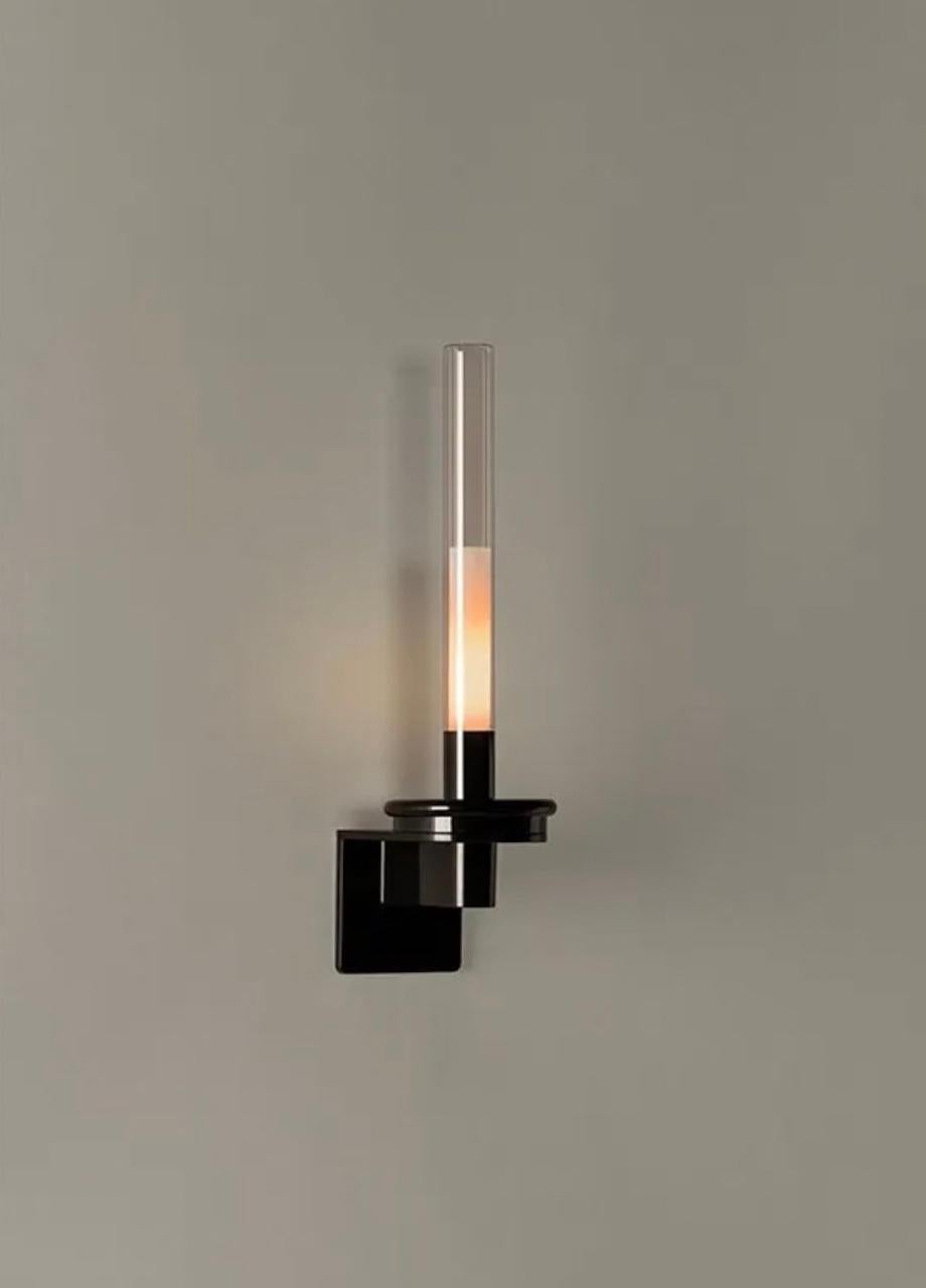 This wall lamp has a circular metal base finished in shiny black and a cylindrical glass shade that diffuses a warm light. Keeping the inspiration of its designers based on the traditional candle, the Sylvestrina wall lamp provides an ambient light