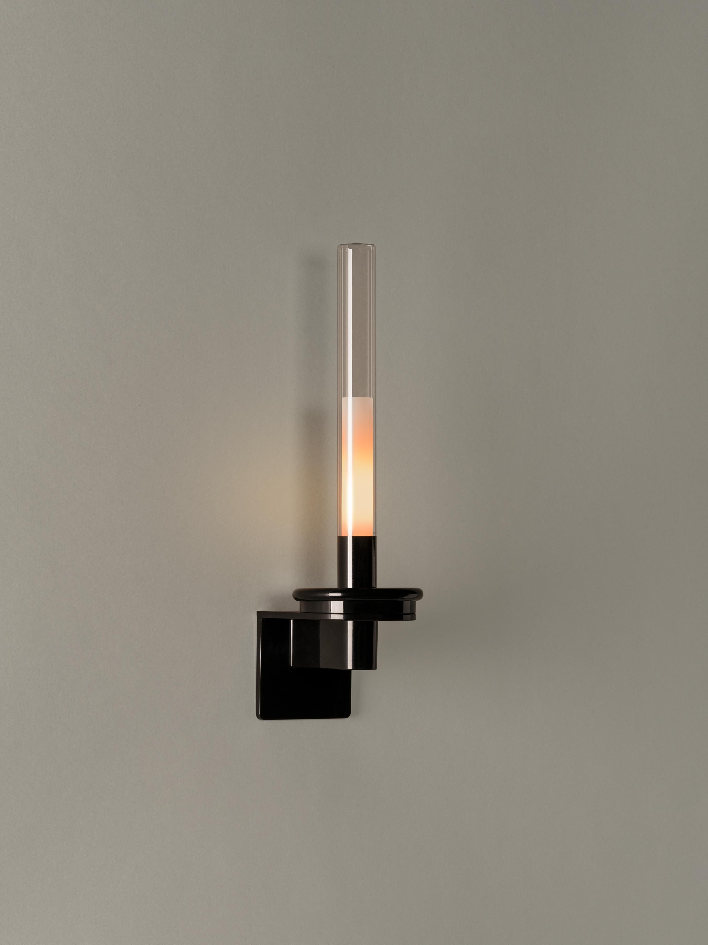 Sylvestrina wall lamp by Enric Sòria, Jordi Garcés
Dimensions: D 12.7 x W 15.1 x H 46 cm
Materials: Metal, glass.

This wall lamp has a circular metal base finished in shiny black and a cylindrical glass shade that diffuses a warm light. Keeping