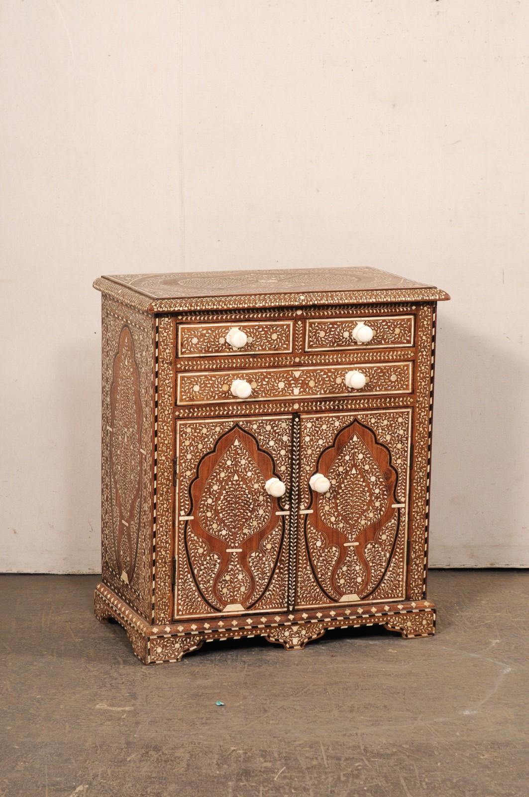 A petite Syrian cabinet, with ornately decorated bone inlay exterior, from the mid 20th century. This mid-century cabinet from Syria is adorn with intricate foliate and floral inlays in born throughout, giving this piece a wonderful sense of texture