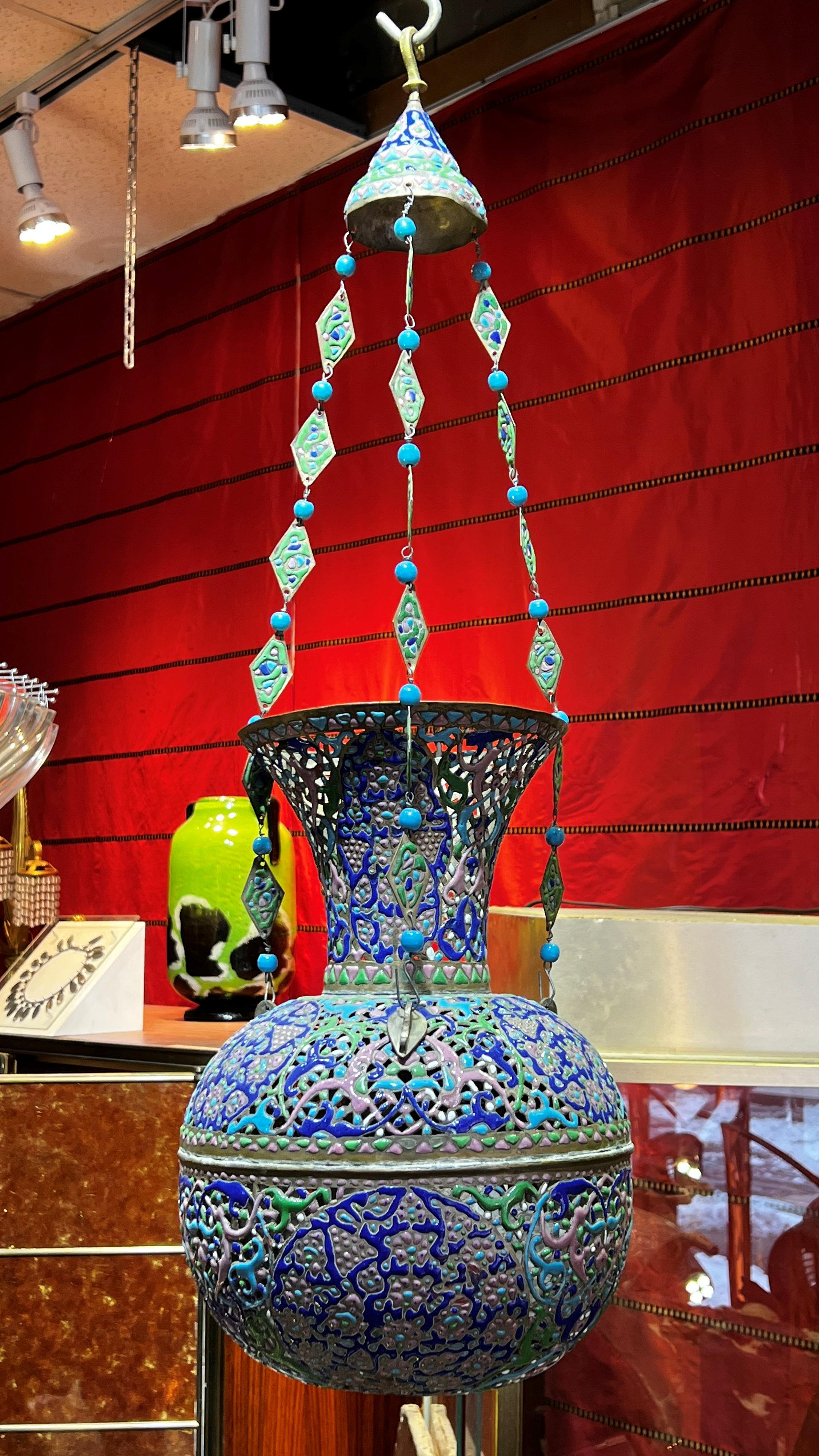 Notre belle lampe mosquée en cuivre se distingue par sa forme réticulée et ses ravissants motifs d'arabesques émaillées en bleu, vert lime et rose, avec ses chaînes de suspension originales formées de perles bleues et de métal émaillé en forme de