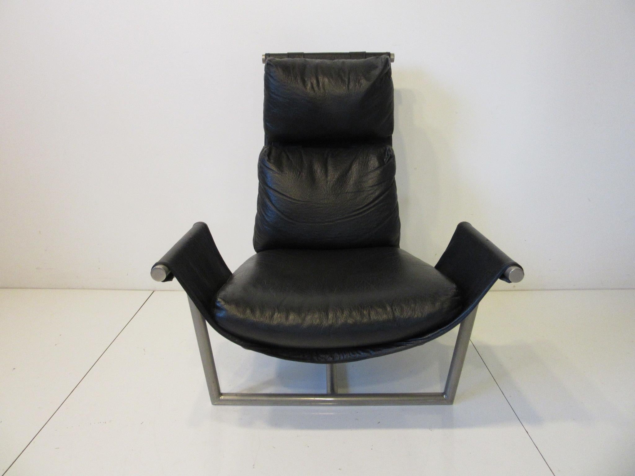 T backed lounge chair with black leatherette cushions sitting on a metal tubed frame in a pewter tone. Very comfortable with puffy cushions on a matching black material sling styled in the manner of Laverne and the Katovolos chair. Retains the