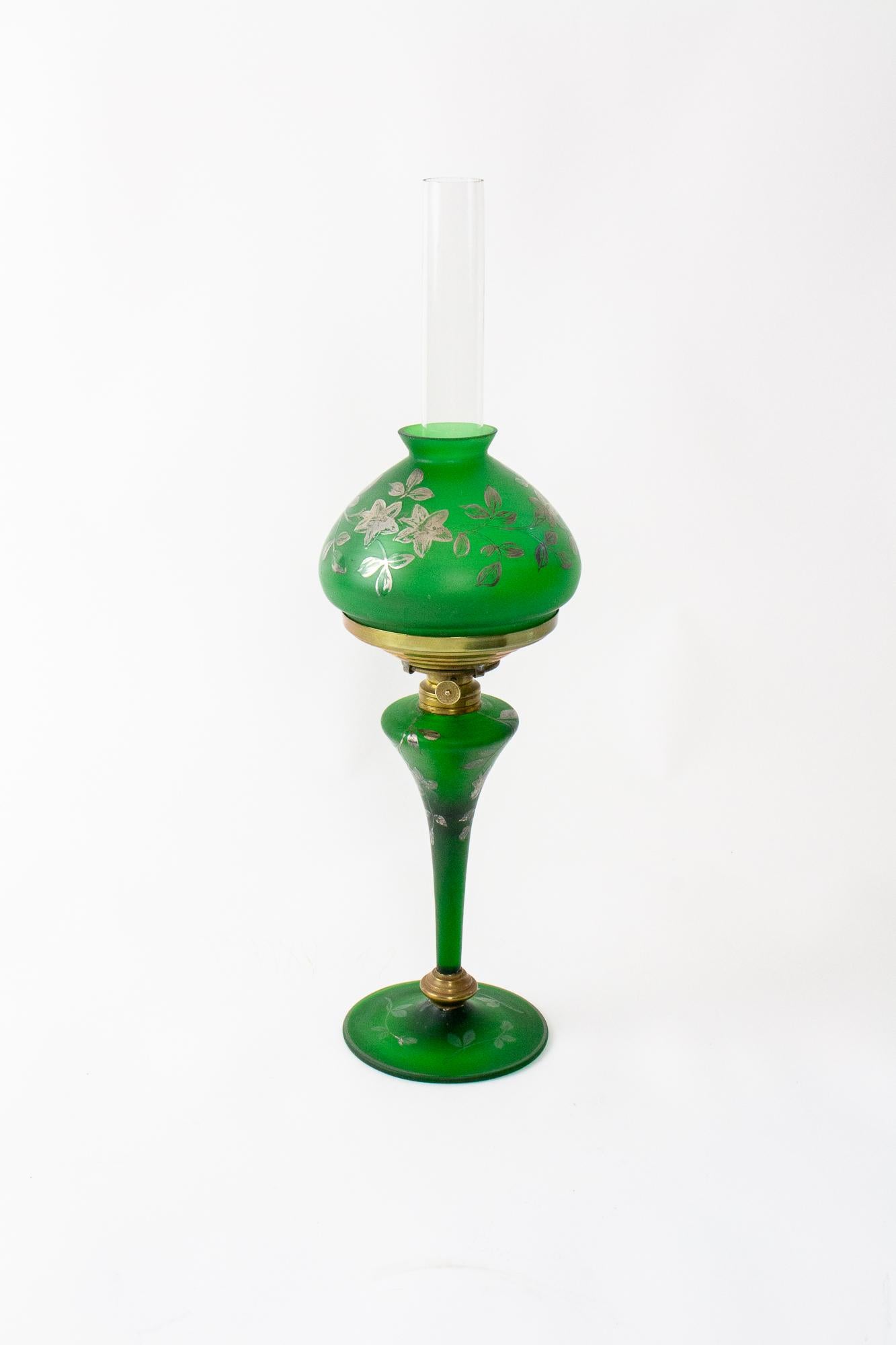 Green art glass lamp. Burner is marked “Manhattan Brass Co.” Body and shade are a frosted green blown art glass with hand painted silver flowers. Sleek Art nouveau profile, the glass base tapers to a round base. The matching shade is a domed shape.