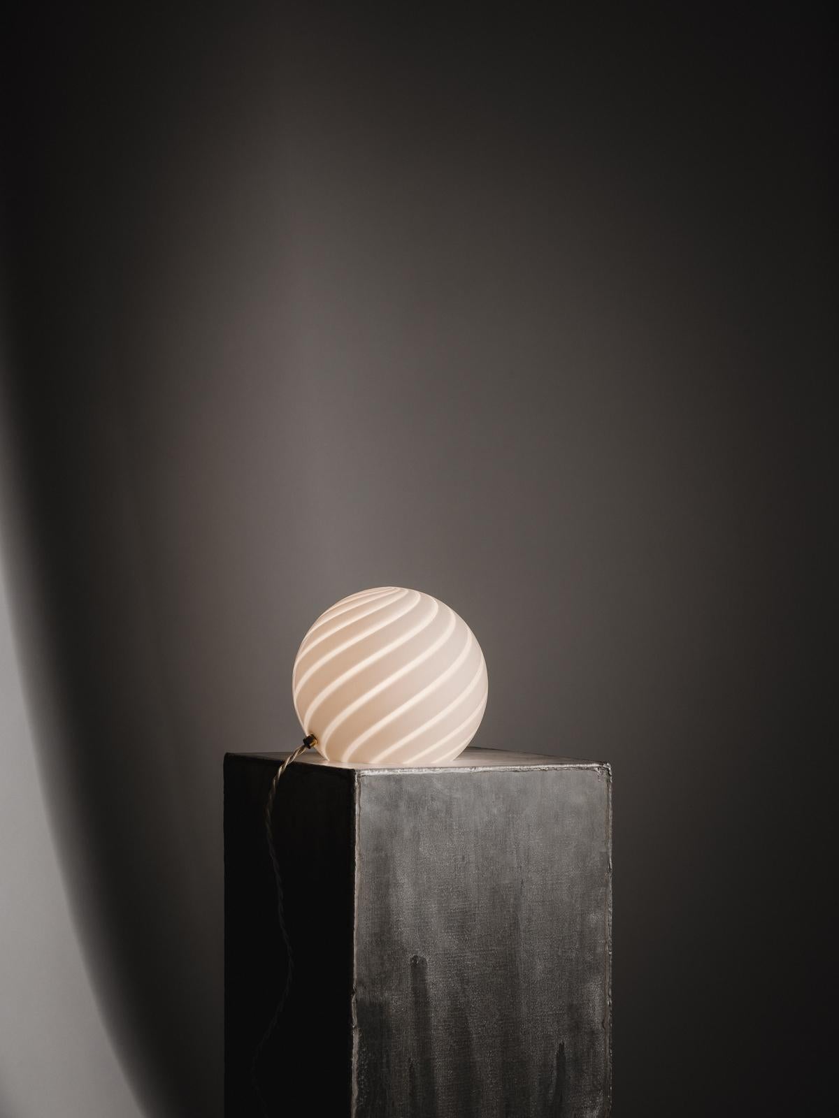 Sphere-shaped table lamp crafted from mouth-blown opaline glass. The design
features a swirl pattern obtained using an original 1970s mould, specifically picked to give the lamp a clean expression. The piece includes hand-casted brass