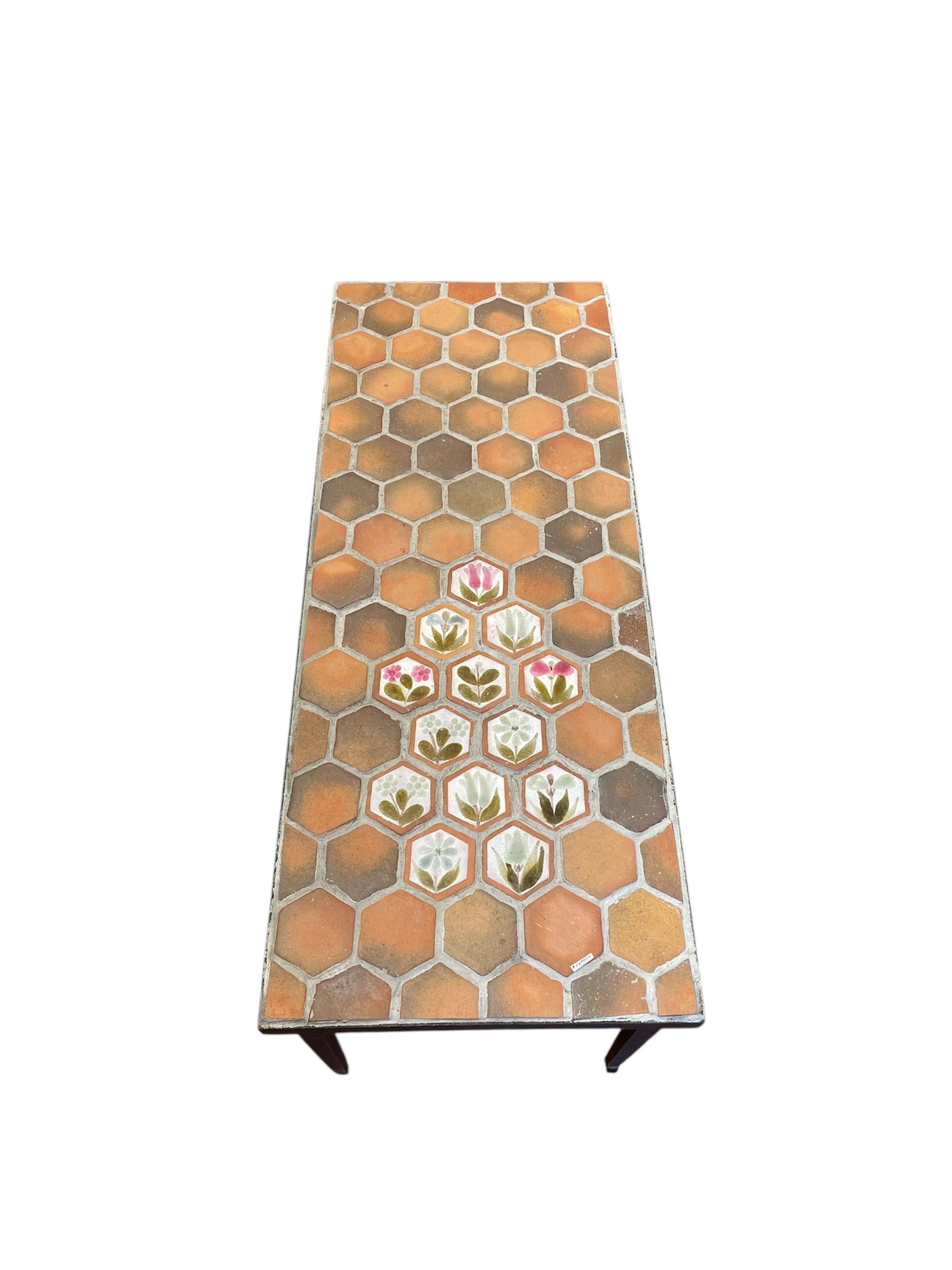 This coffee table features a top adorned with hexagonal terracotta tiles, typical of Roger Capron's work, along with a set of decorative glazed tiles with floral motifs. The base is simple and slender, highlighting the ceramic top. The overall