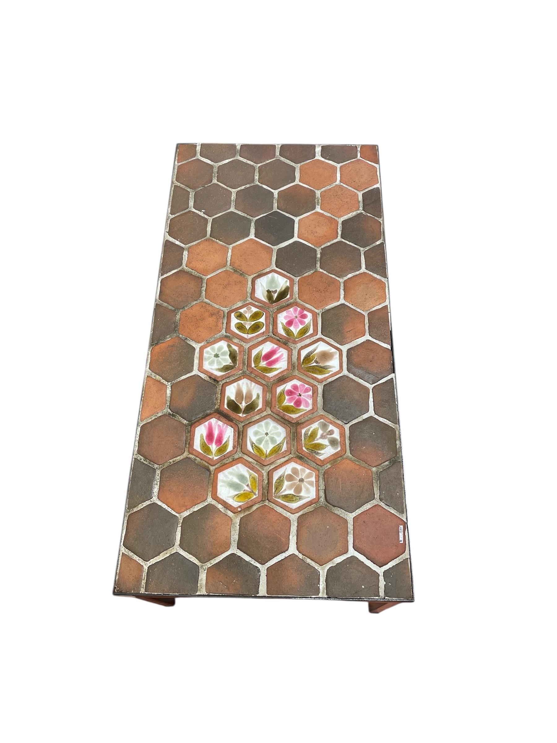 This coffee table features a top adorned with hexagonal terracotta tiles, typical of Roger Capron's work, along with a set of decorative glazed tiles with floral motifs. The base is simple and slender, highlighting the ceramic top. The overall