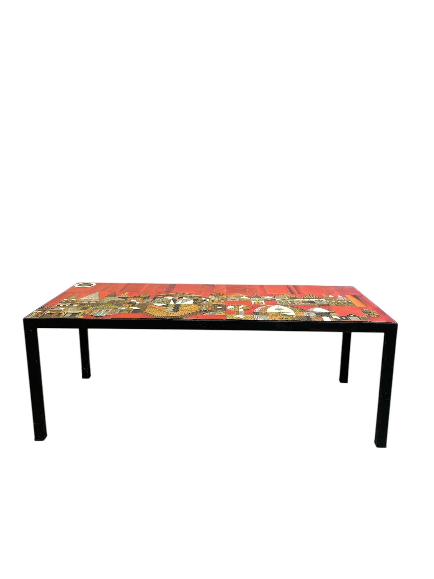 Table basse attribuée à Roger Capron, avec un plateau en céramique émaillée rouge décoré d'une scène de village stylisée. La pièce présente une composition graphique dynamique, mêlant des teintes vibrantes et des motifs architecturaux