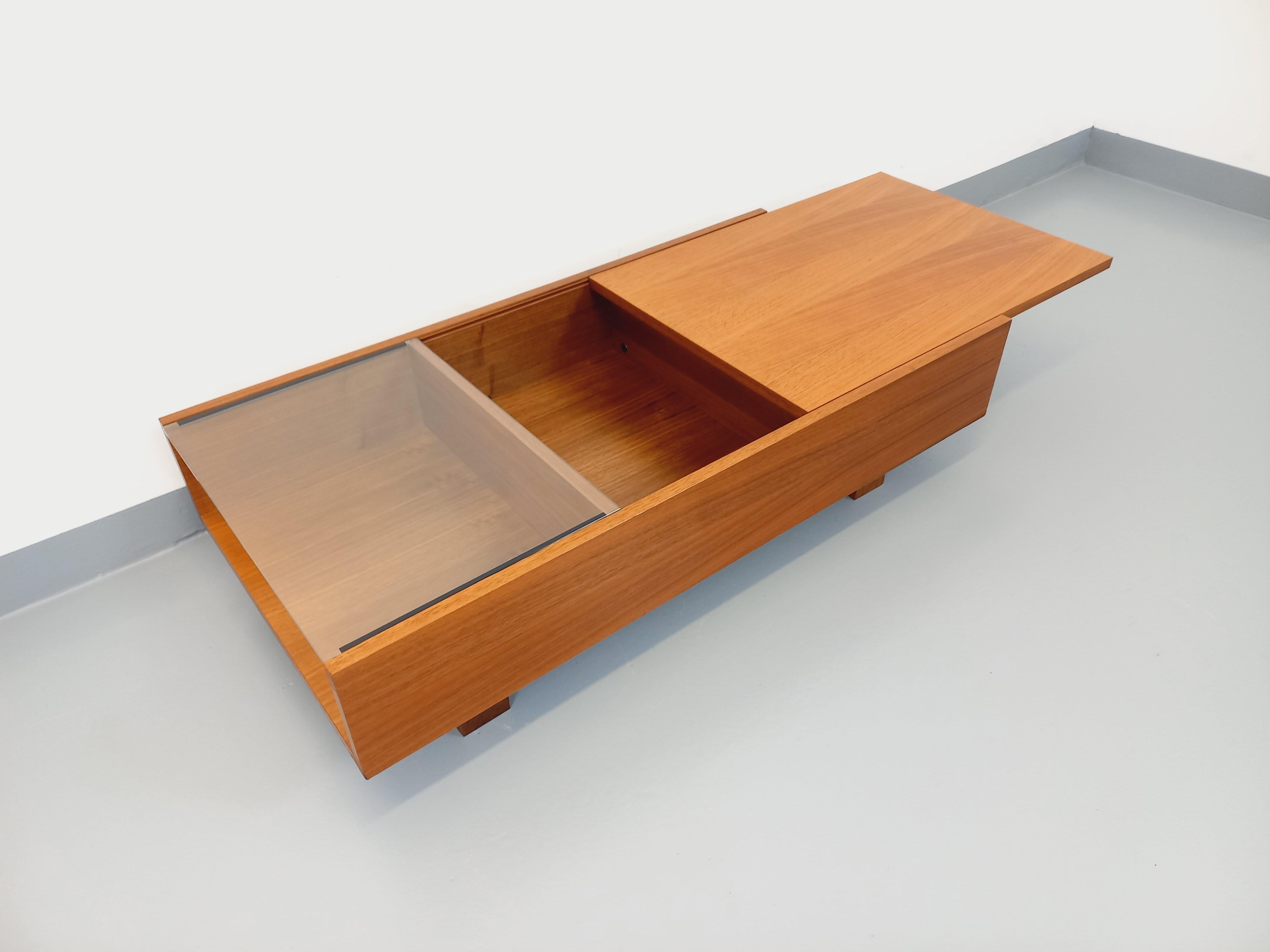 Table basse rectangulaire moderniste vintage des années 60 70, très beau design en teck et verre transparent, 2 niches ouvertes extérieures, et 1 coffre intérieur grâce à un plateau coulissant. Très bon état général avec quelques signes du temps qui