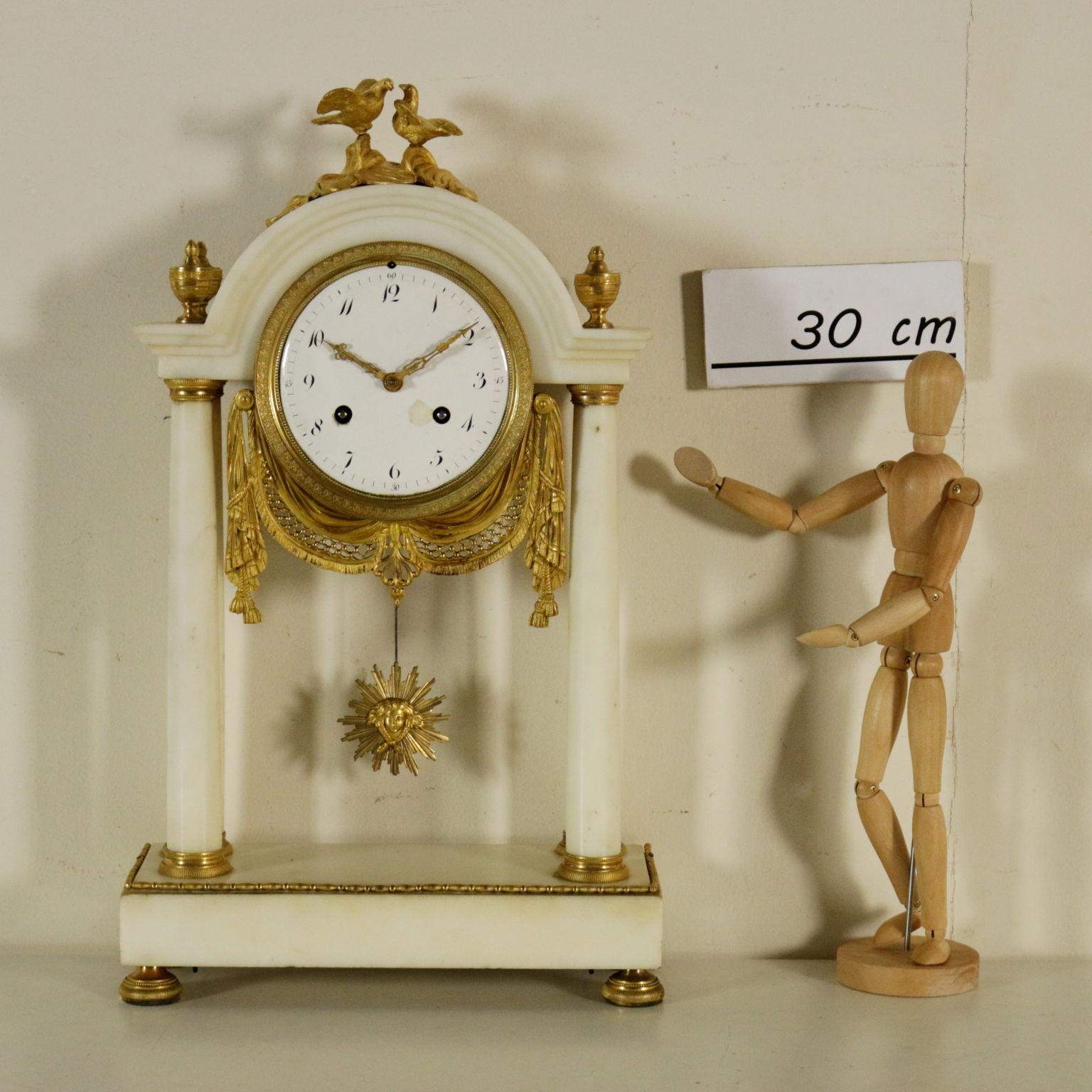 A table clock, white marble and gilded bronze, bulin treated. Glazed metal face with Arabian numbers enclosed in a bronze part and decorated with a large drape. The clock hands are made of finely perforated metal. A pair of doves on the top on sea