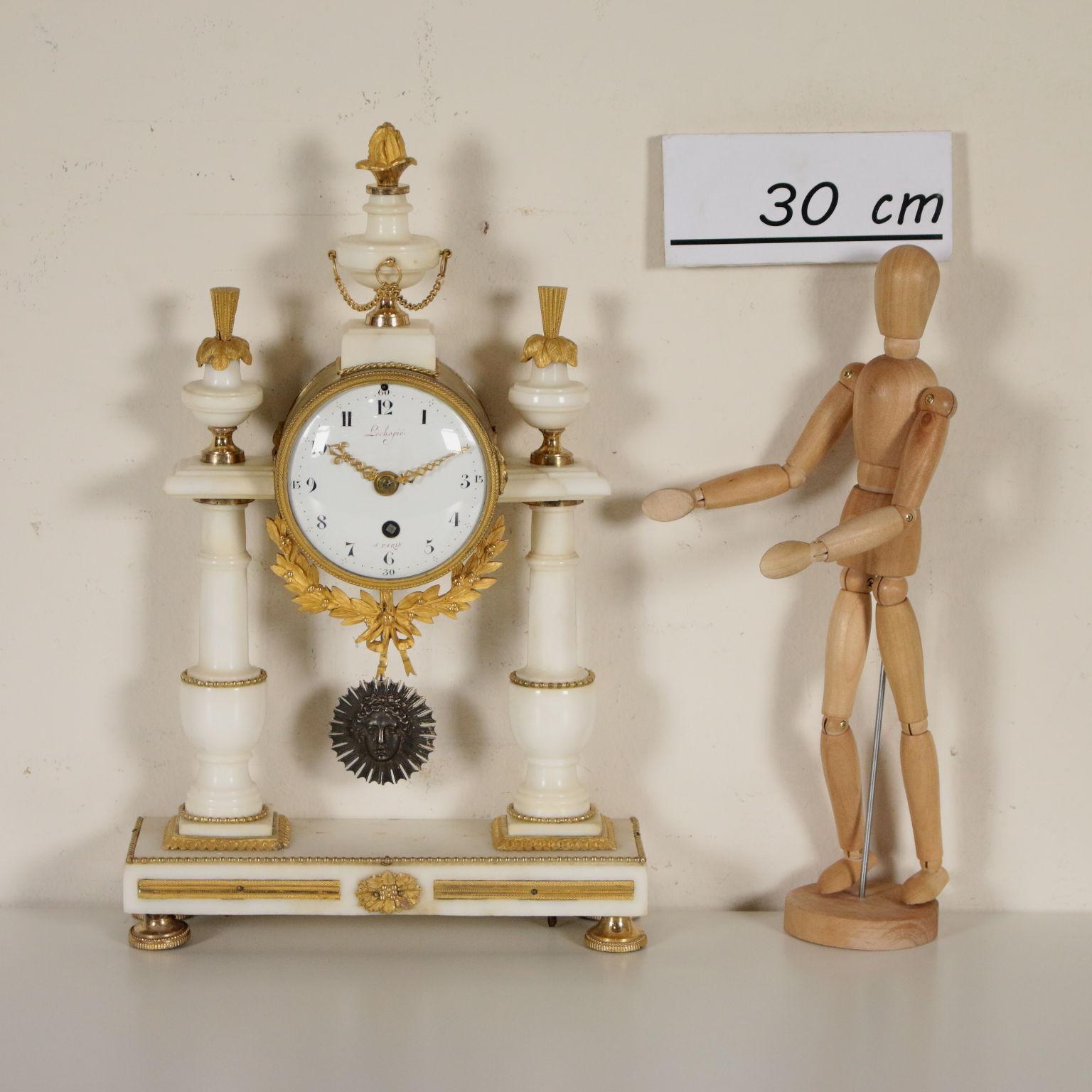 Une horloge de table en marbre blanc avec des détails en bronze doré. Sous-sol rectangulaire avec deux colonnes tournées et une horloge cylindrique entre elles. Le marbre blanc est agrémenté de détails en bronze doré, d'encadrements, de flammes et