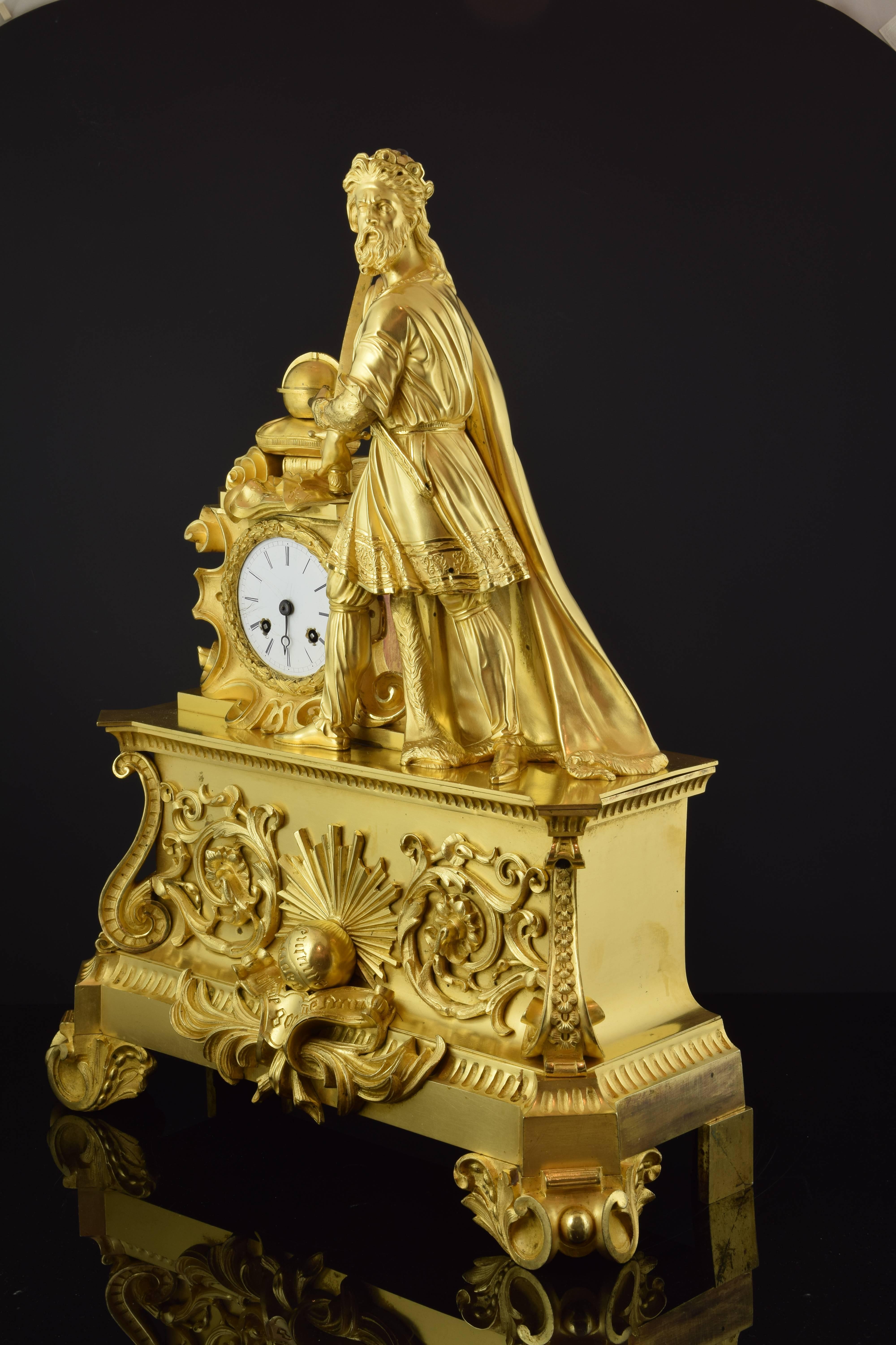 Table clock made of gilded bronze with a powerful base, enhanced with legs decorated with vegetal and architectural motifs of classicist inspiration and a series of details on the front (volutes in the chamfered corners, volutes and a center with a