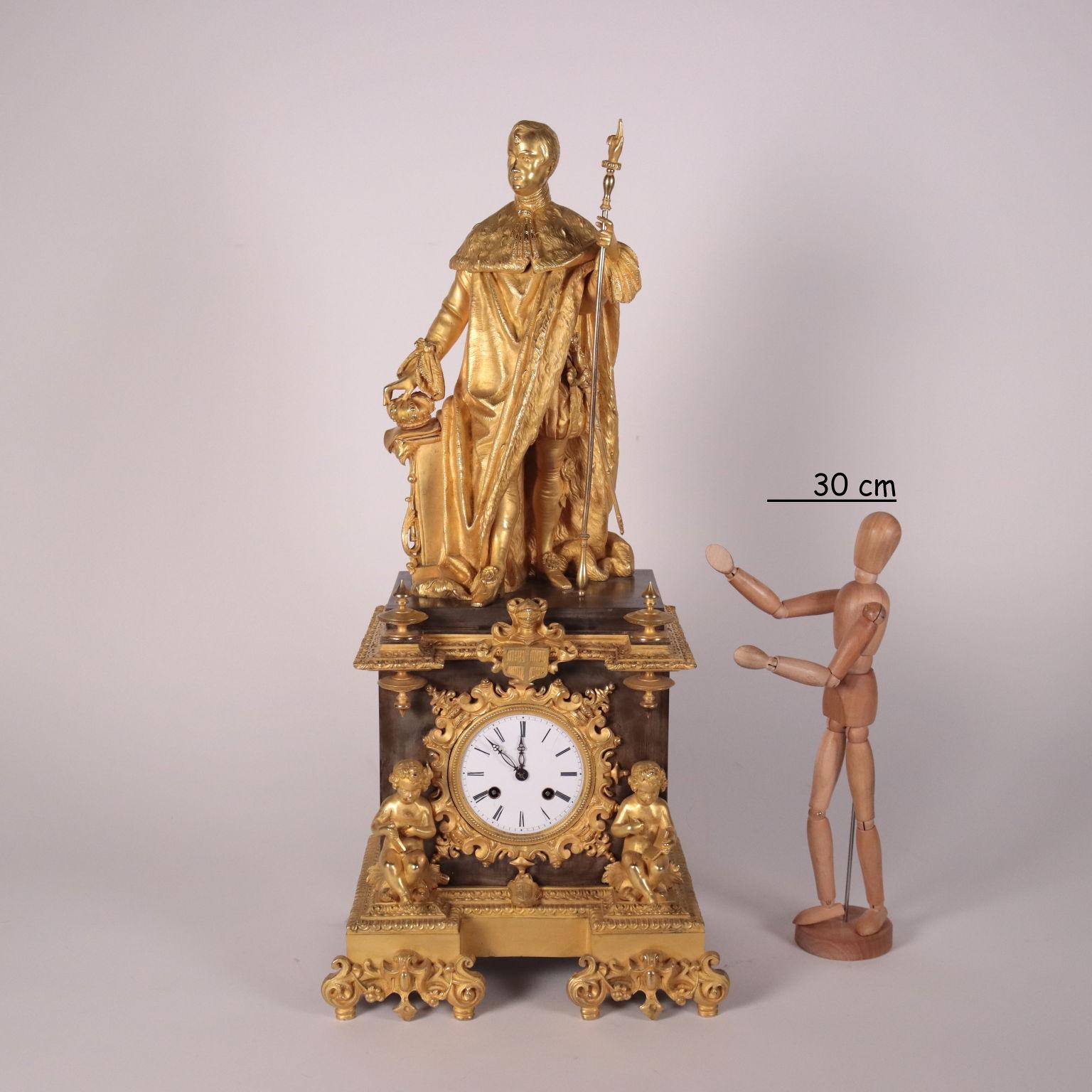 Table clock in bronze with dark and gilded patina. Two cherubs seated and intent on writing flank the clock. The enameled metal display with Roman numeral hours is enclosed within a gilt bronze wreath. At the top there is a statue of an emperor; the