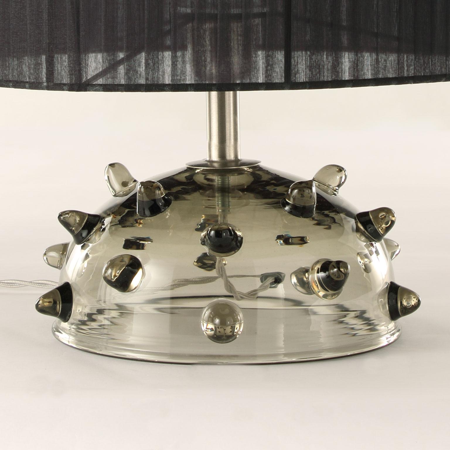 Table lamp in grey artistic glass with clear “bugne” (particular application in glass on the base), black organza handcrafted lampshade.

Size: 35 length x 41 height

The handcrafted soul of our lighting products is reinvented every time we start a