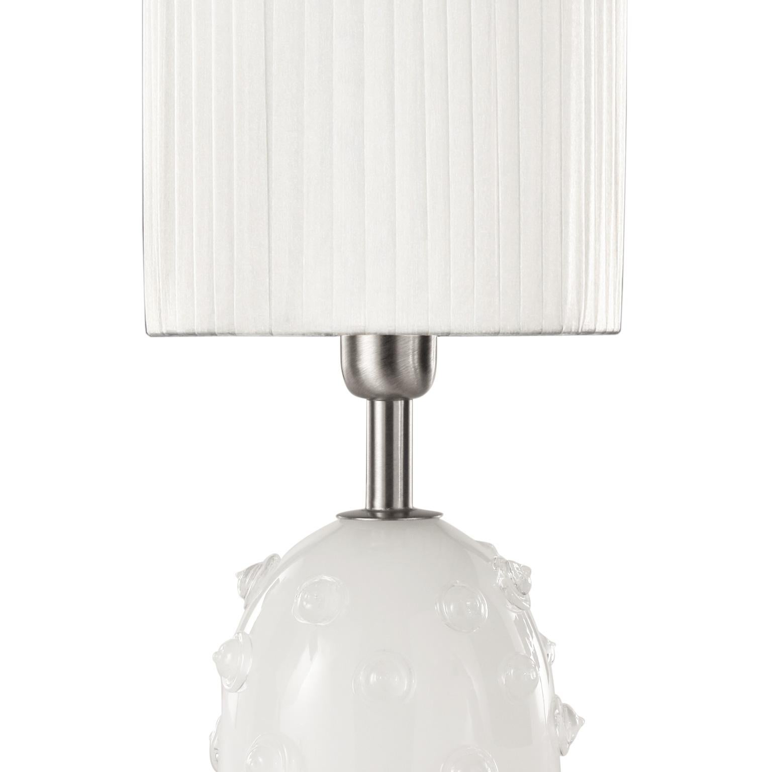 Table lamp in silk artistic Murano glass with clear “borchie” (particular application in glass on the base), grey organza lampshade. Nickel fixture.
The handcrafted soul of our lighting products is reinvented every time we start a new project, when