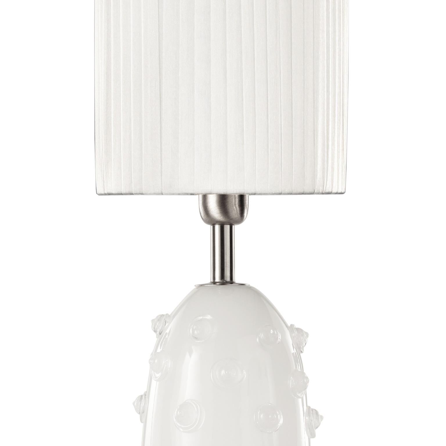 Table lamp in silk artistic Murano glass with clear “borchie” (particular application in glass on the base), grey organza lampshade. Nickel fixture.
The handcrafted soul of our lighting products is reinvented every time we start a new project, when