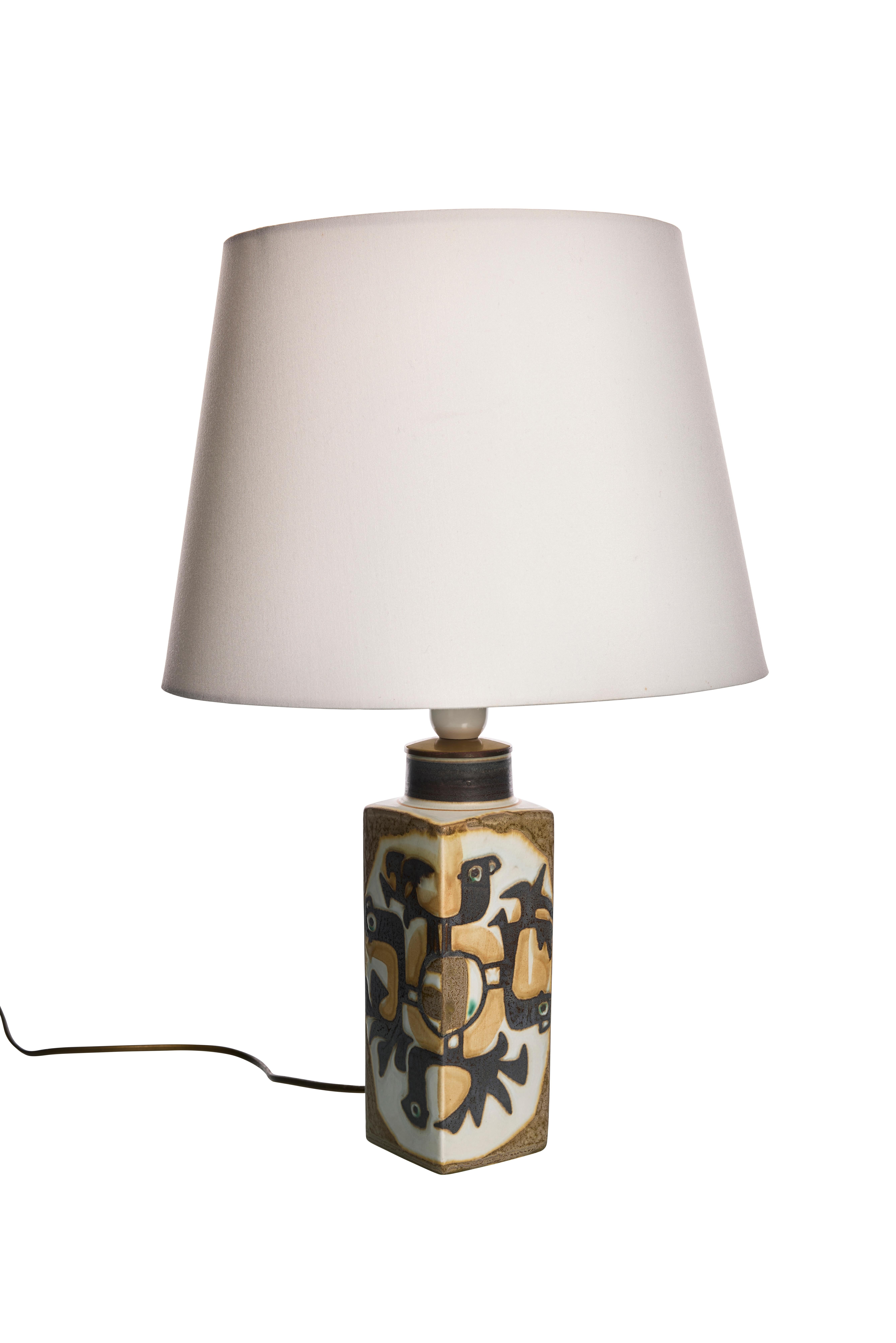 Questa lampada da tavolo, disegnata da Johanne Gerber e Nils Thorsson per Royal Copenhagen, presenta una base rettangolare in ceramica ornata da un disegno astratto dai toni della terra che comprende forme nere e marroni, forse simili a uccelli. Il