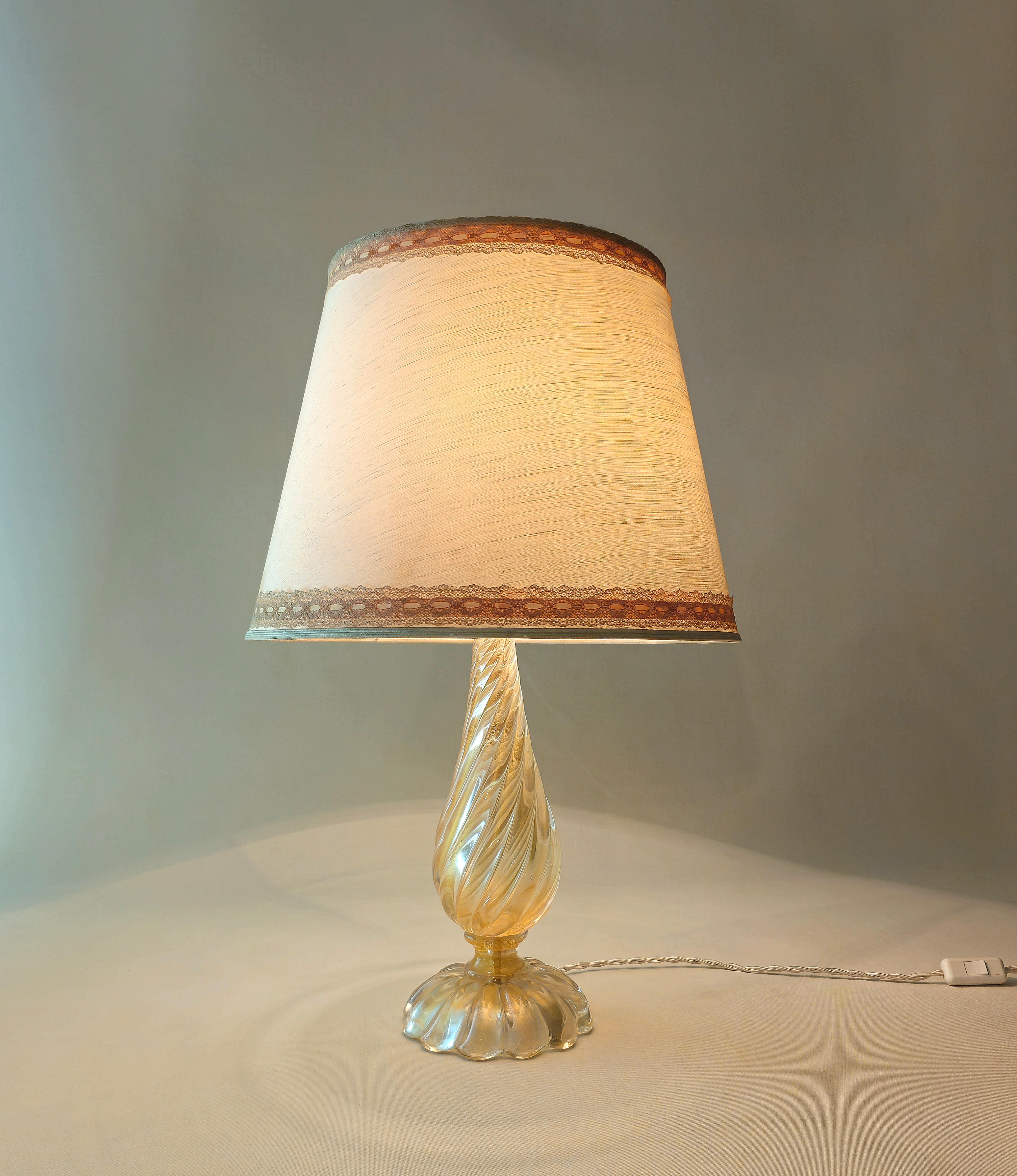 Barovier&Toso, Italie Années 1970.
Grande lampe de table E27 à 1 lumière en verre de Murano transparent et nervuré or, tige en forme de goutte, base circulaire nervurée et abat-jour en tissu ivoire avec bordure brodée.

Note : Nous essayons d'offrir