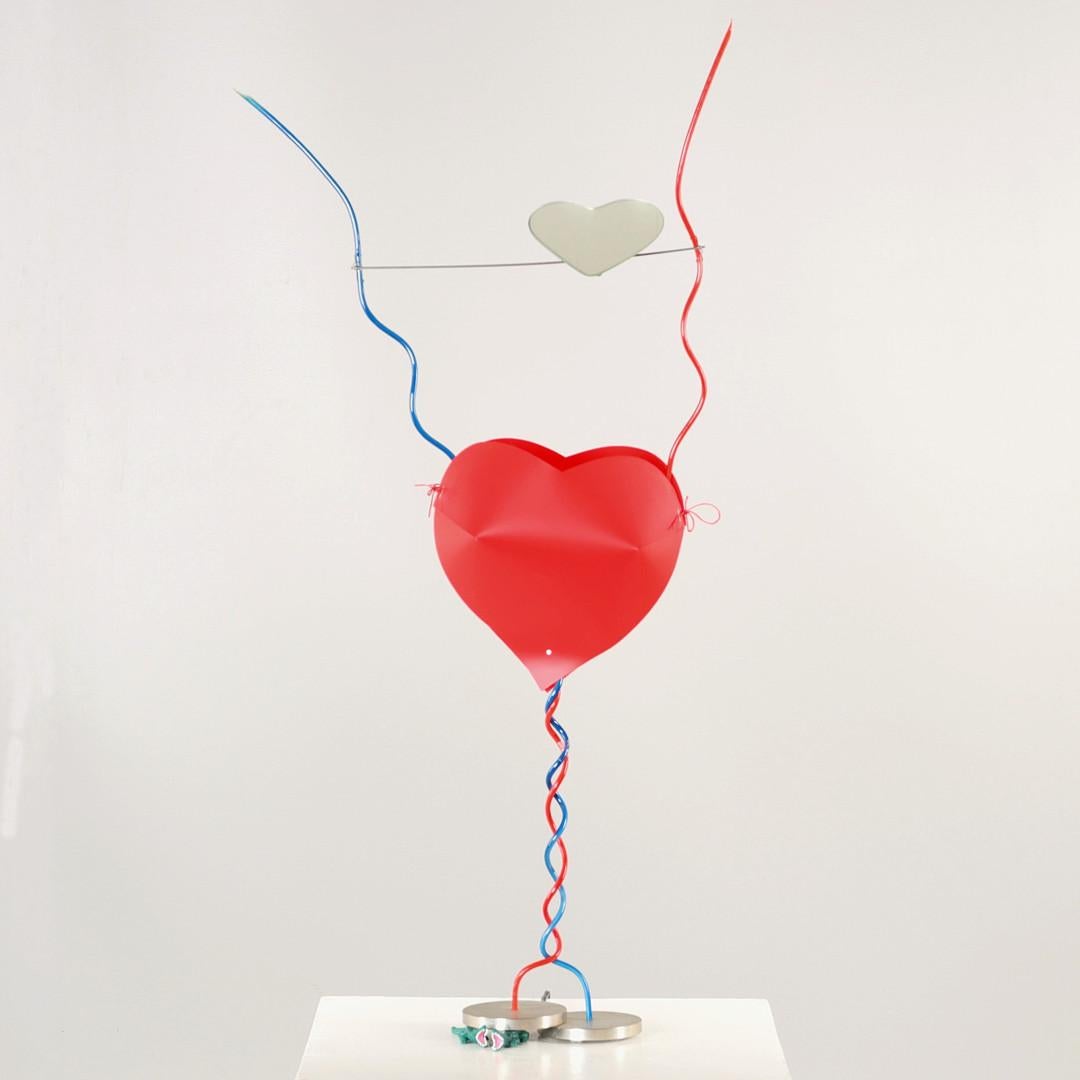 “One from the heart” table lamp, a wandering heart on two metal bases, one of which rests on two plastic crocodiles. Metal leg stand with plastic housing in red and blue. In the middle the light source is surrounded by a plastic heart, above the