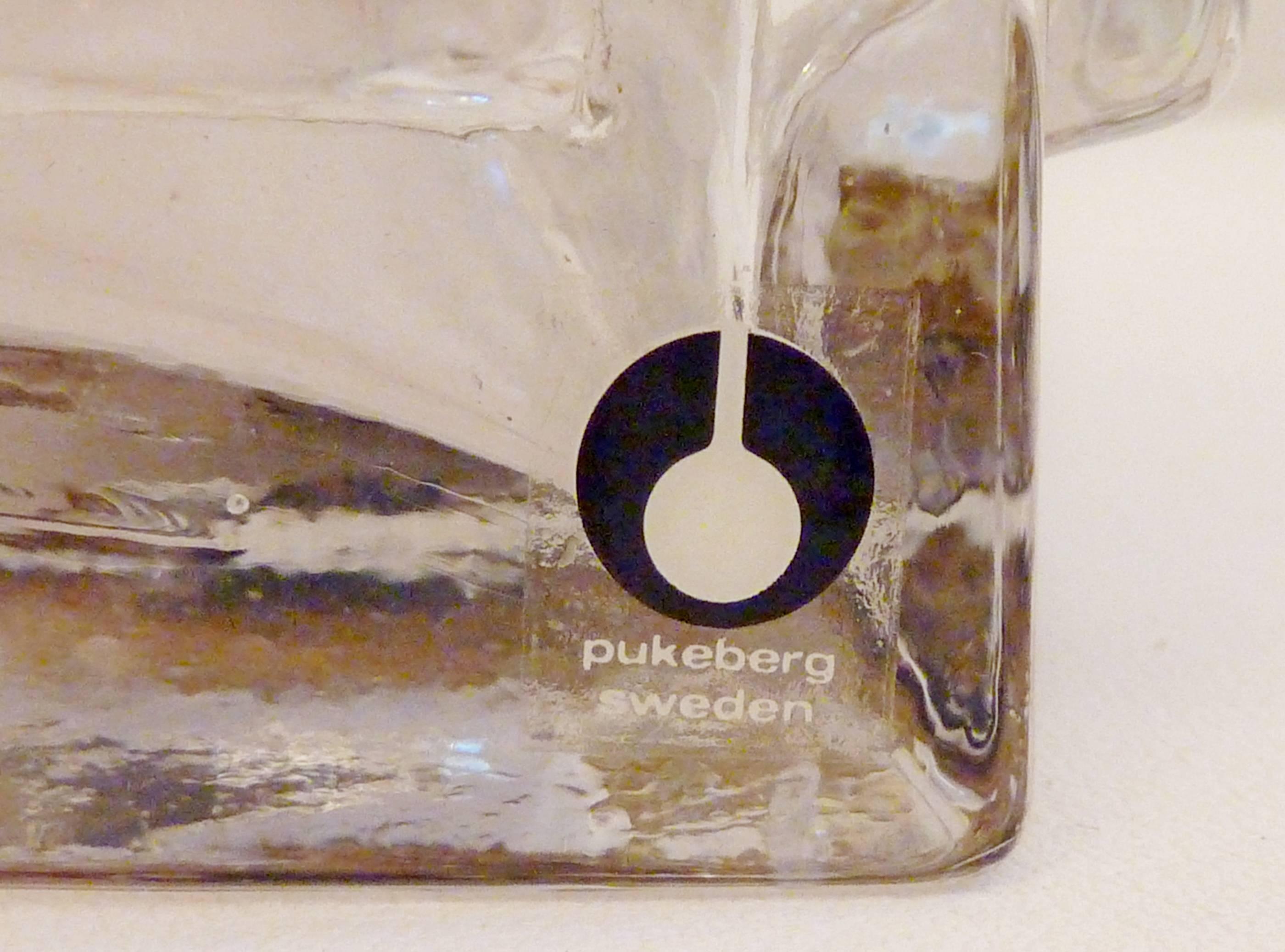 Lampe de table en verre transparent du fabricant suédois Pukeberg avec autocollant original sur la base. Livré avec un abat-jour blanc doublé d'argent qui reflète bien la lumière.