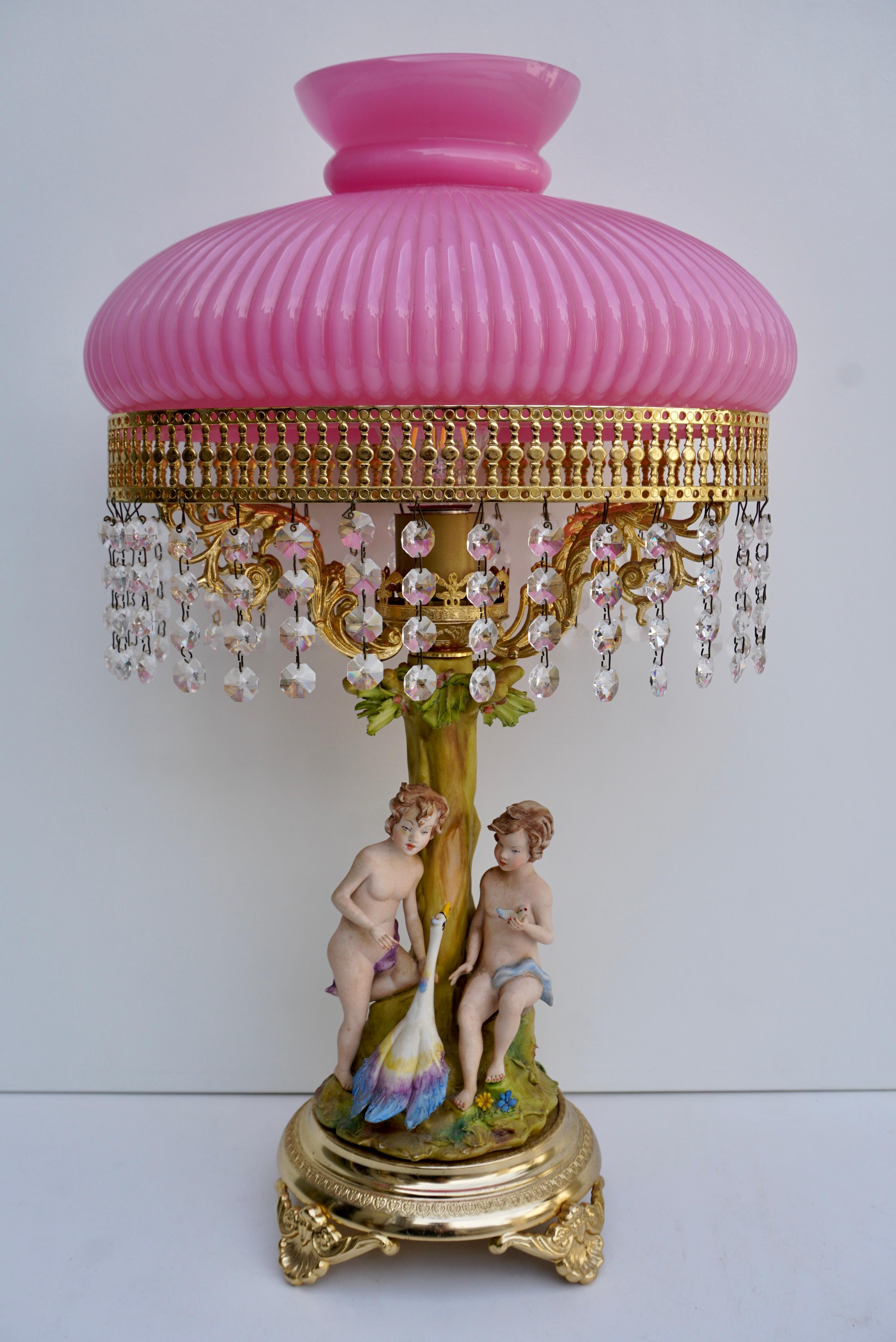 Italian table lamp in porcelain and glass.
Two children with a swan under a tree”. A porcelain statue mounted as a lamp with pink glass shade and crystal bells.