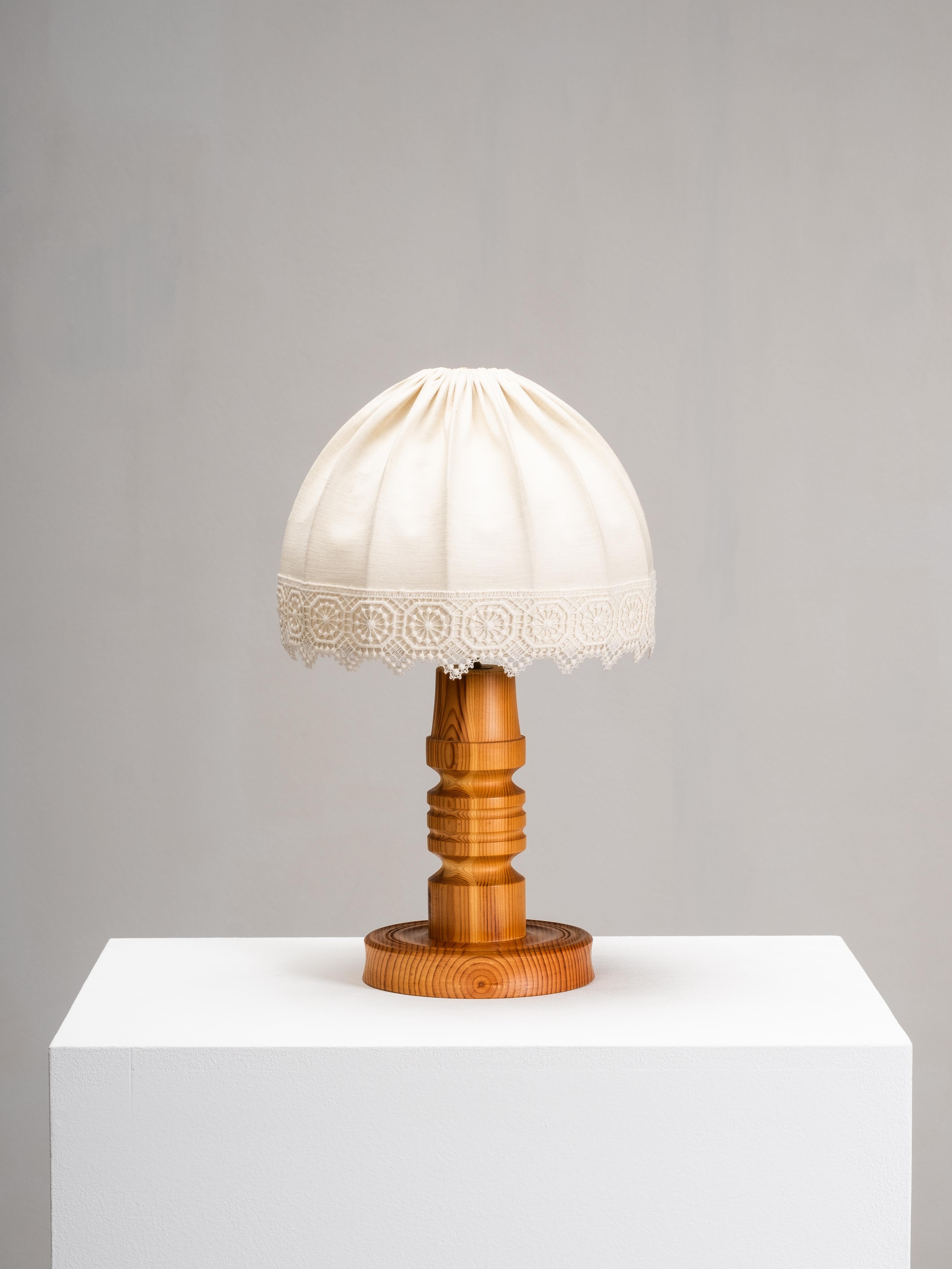 Lampe de table suédoise des années 1970 avec une base en pin tourné et un abat-jour en tissu froncé bordé de dentelle. La base sculpturale montre le grain naturel du pin, avec son ton chaud et ses anneaux de croissance distinctifs, tandis que
