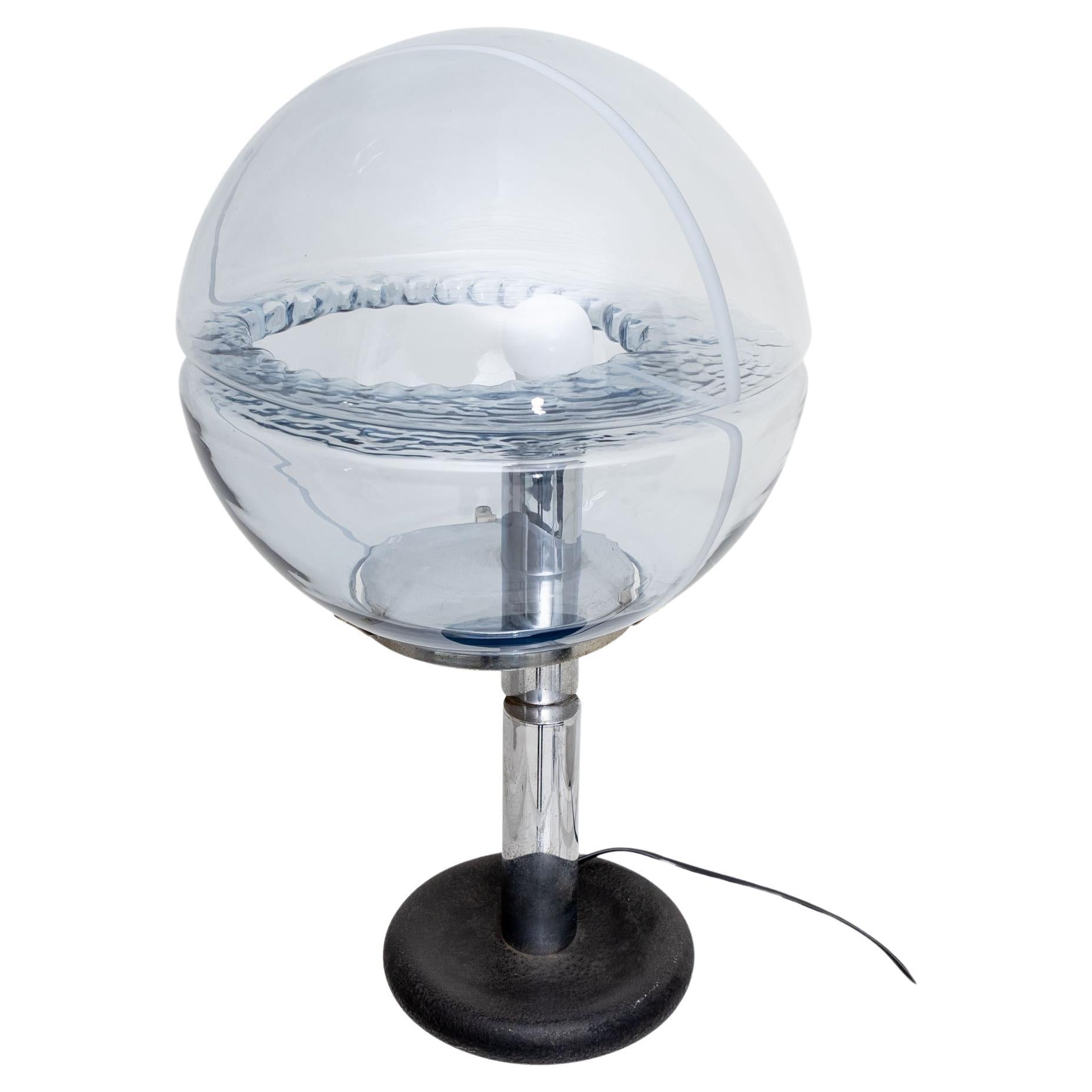 Table lamp with spherical blue tinted glass shade with white stripe decoration and horizontal incision with wavy texture. The large glass shade rests on a tripartite cylindrical stem in chromed metal and a round disc base in iron.