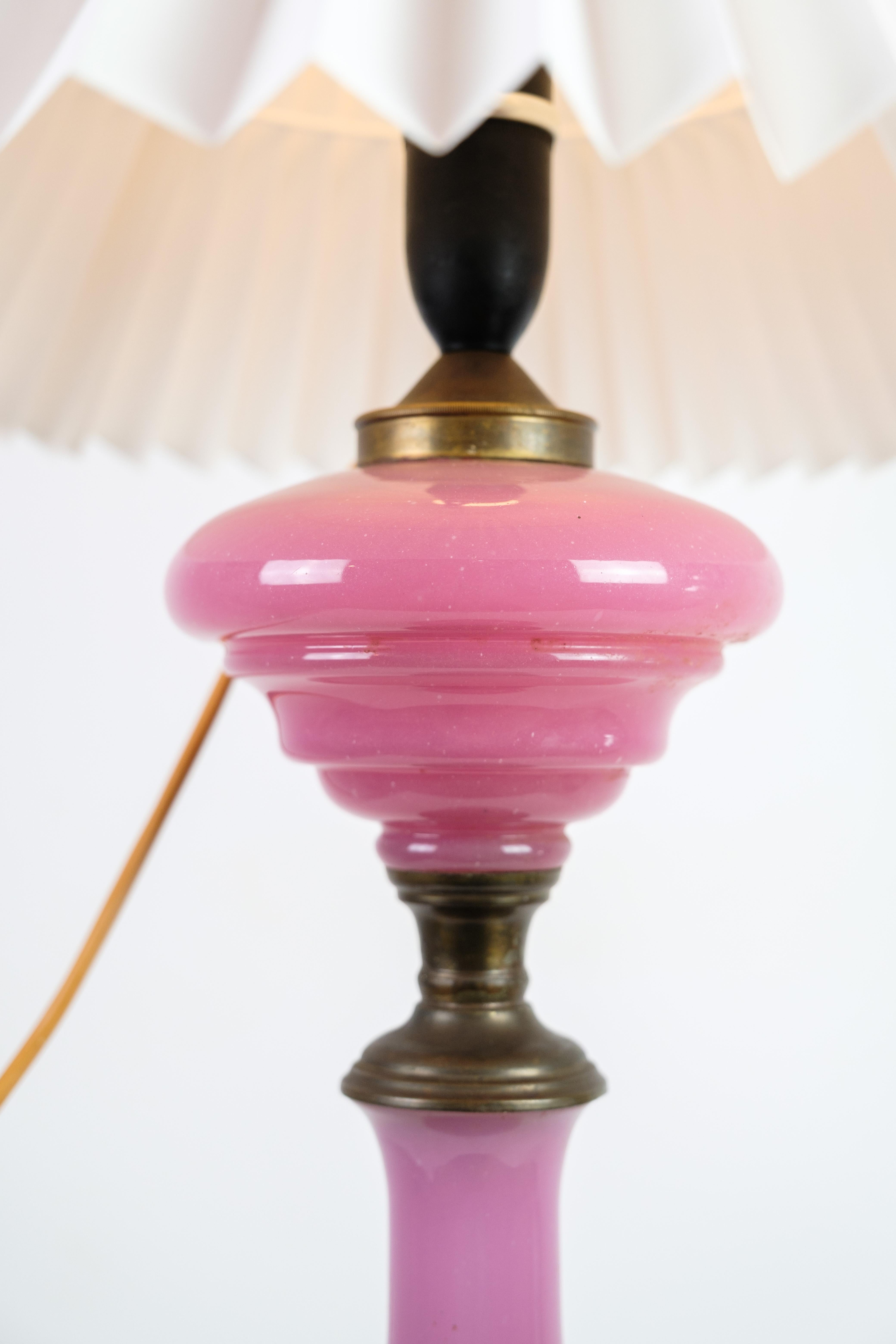 This table lamp is a beautiful example of design from the 1880s. The lamp has a frame of pink opaline glass, which is a type of glass known for its beautiful and characteristic colour. The lamp has a white lampshade. The base is made of brass, which