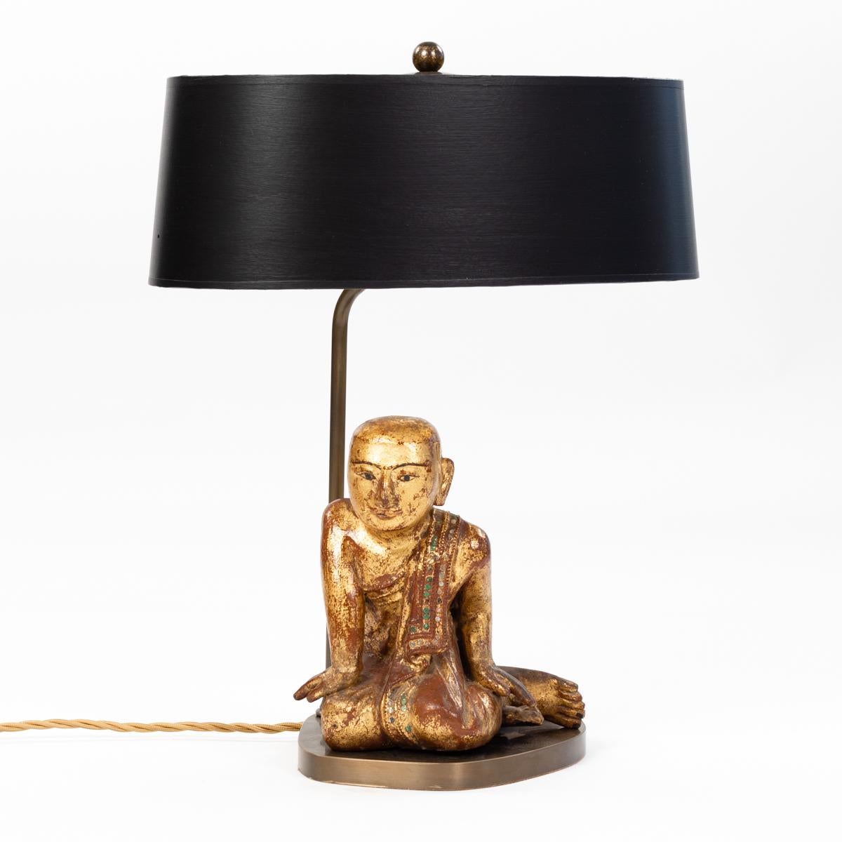 Small table lamp of a sitting priest made of wood with gilded finish. 
The noble object comes from 19th century Burma, the priest has a clear look and pleasing posture. 
The robe has falls backwards over the shoulder casually on the floor.
Borders