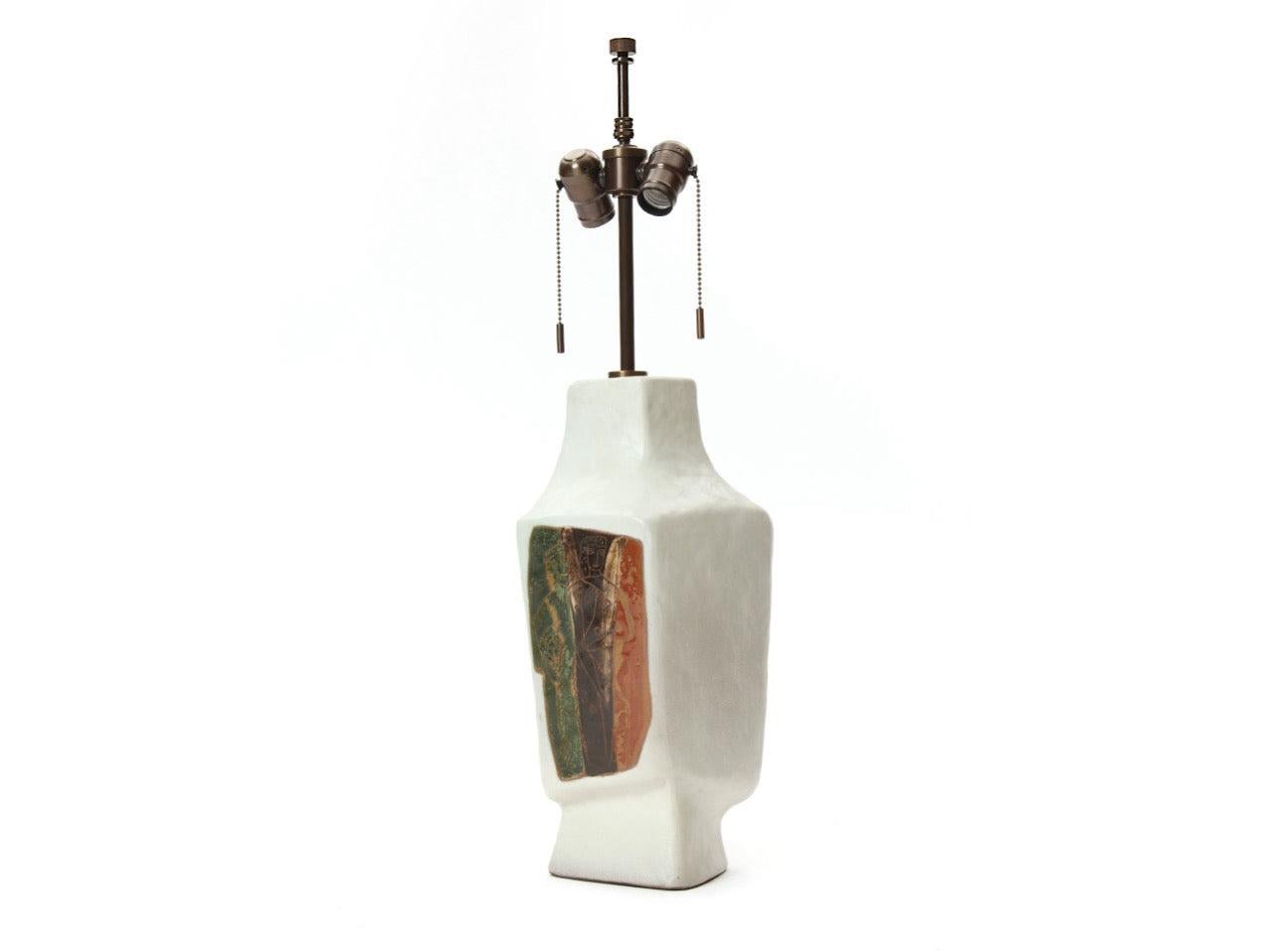 A white ceramic table lamp with two figures etched over green, purple, and orange stripes.