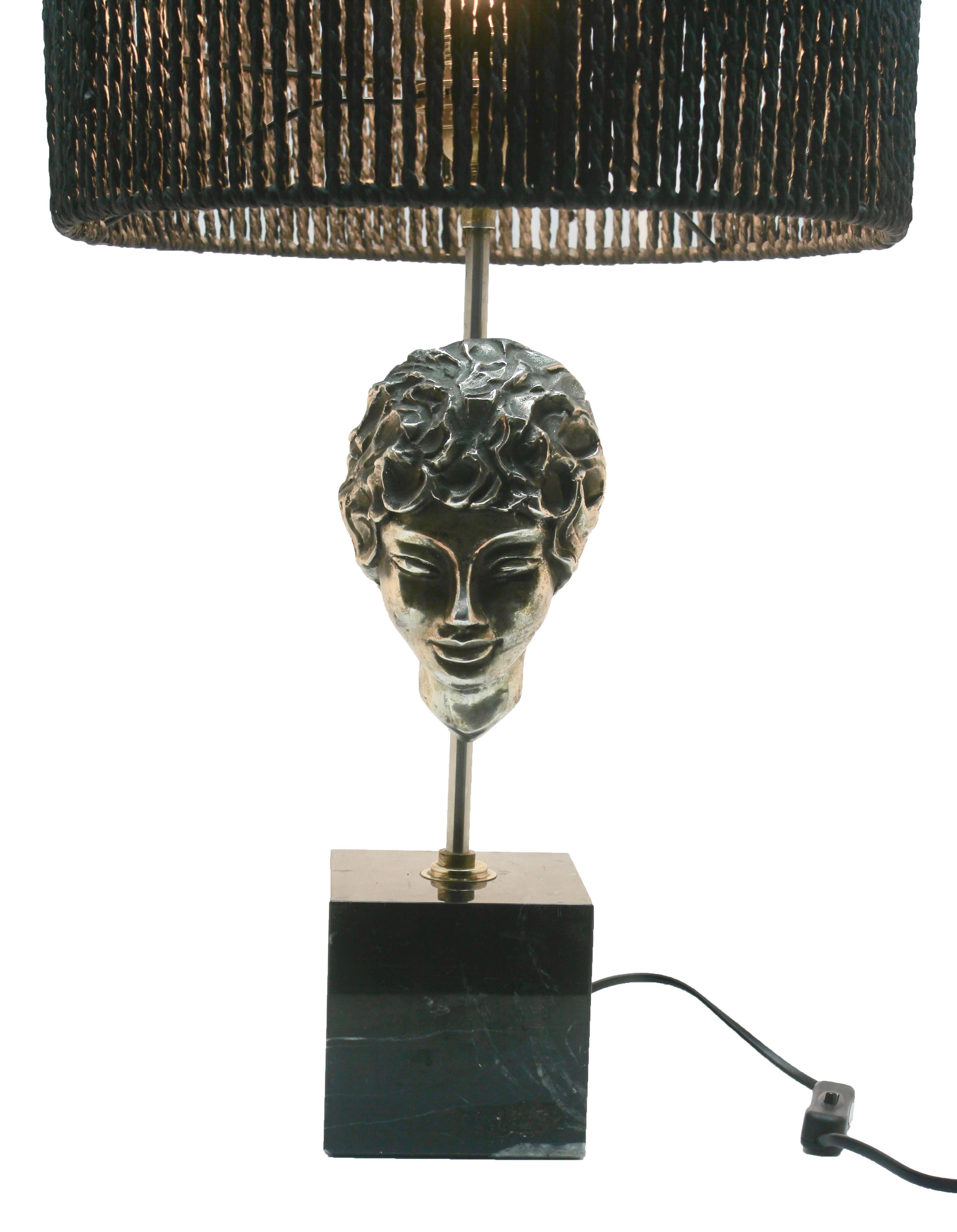 A nicely made table lamp with downlight that catches the facial features in a naturalistic way. 

Polished marble base with a chromed metal pillar supporting a flat-backed masque of a female head in silver-patinated bronze. Probably made during
