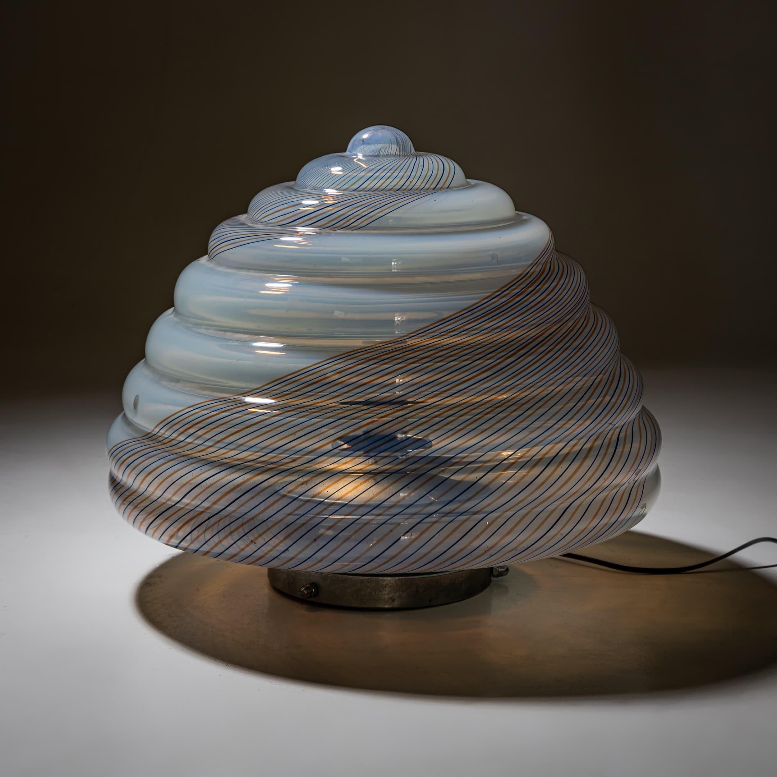 A decorative table lamp featuring a beehive-shaped glass shade with spiral ribbing. The translucent glass is interwoven with fine colored stripes, creating a dynamic, almost organic appearance. Mounted on a circular metal base.