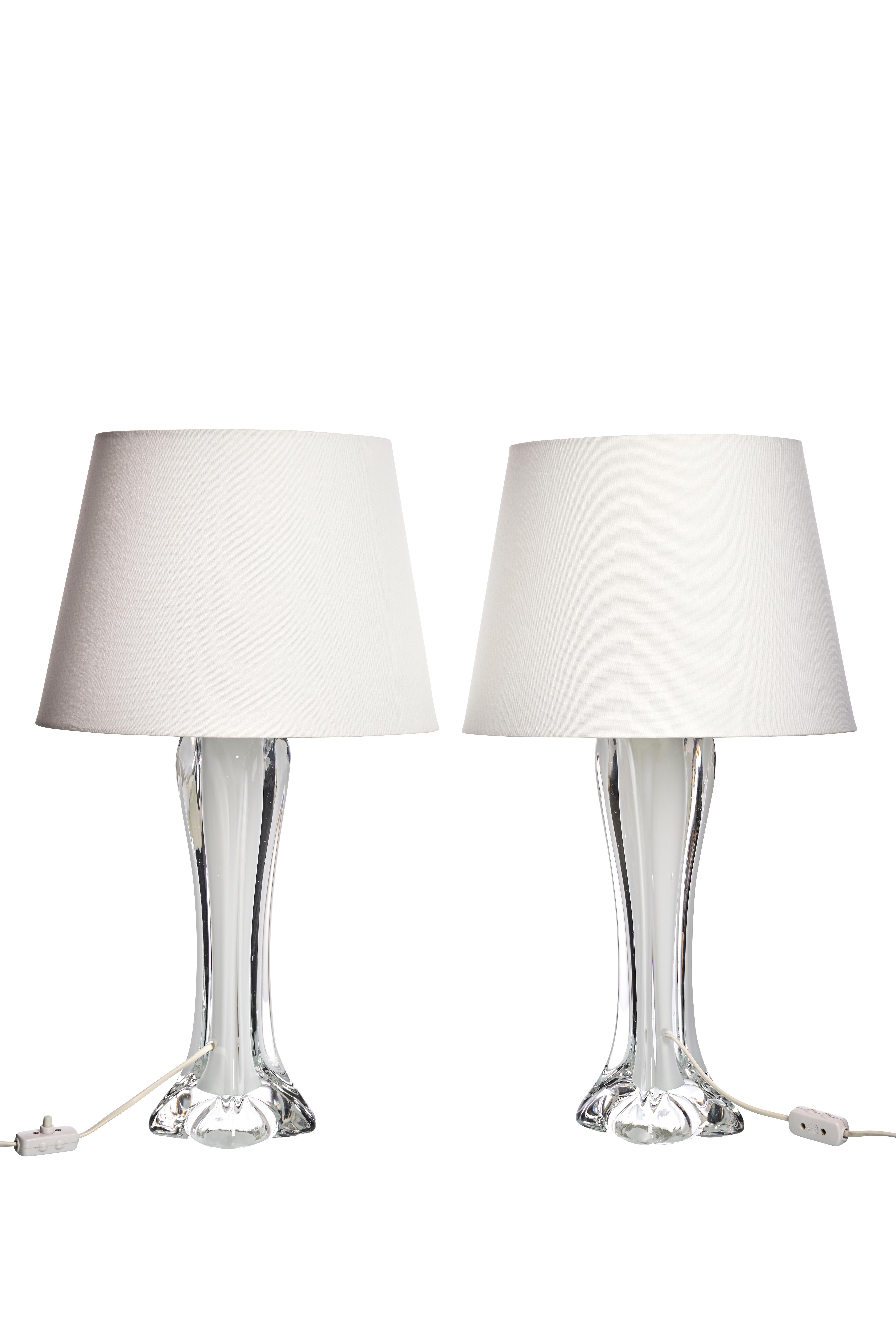Este conjunto de dos lámparas de sobremesa de Paul Kedelv para Flygfors, fabricadas en la década de 1960, presenta un diseño de cristal transparente, elegante y liso. Cada lámpara tiene una base alta y estrecha con curvas fluidas y una forma