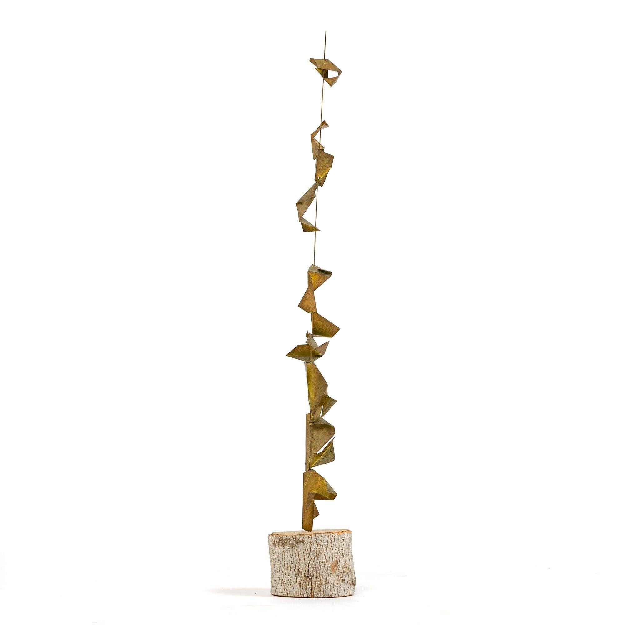 A metal sculpture consisting of a thin rigid wire with folded, fortune-cookie-shape pieces of naturally patinated brass slipped onto the rod and stacked vertically. The rod is set in a round, drum shaped slab of birch retaining its original bark.