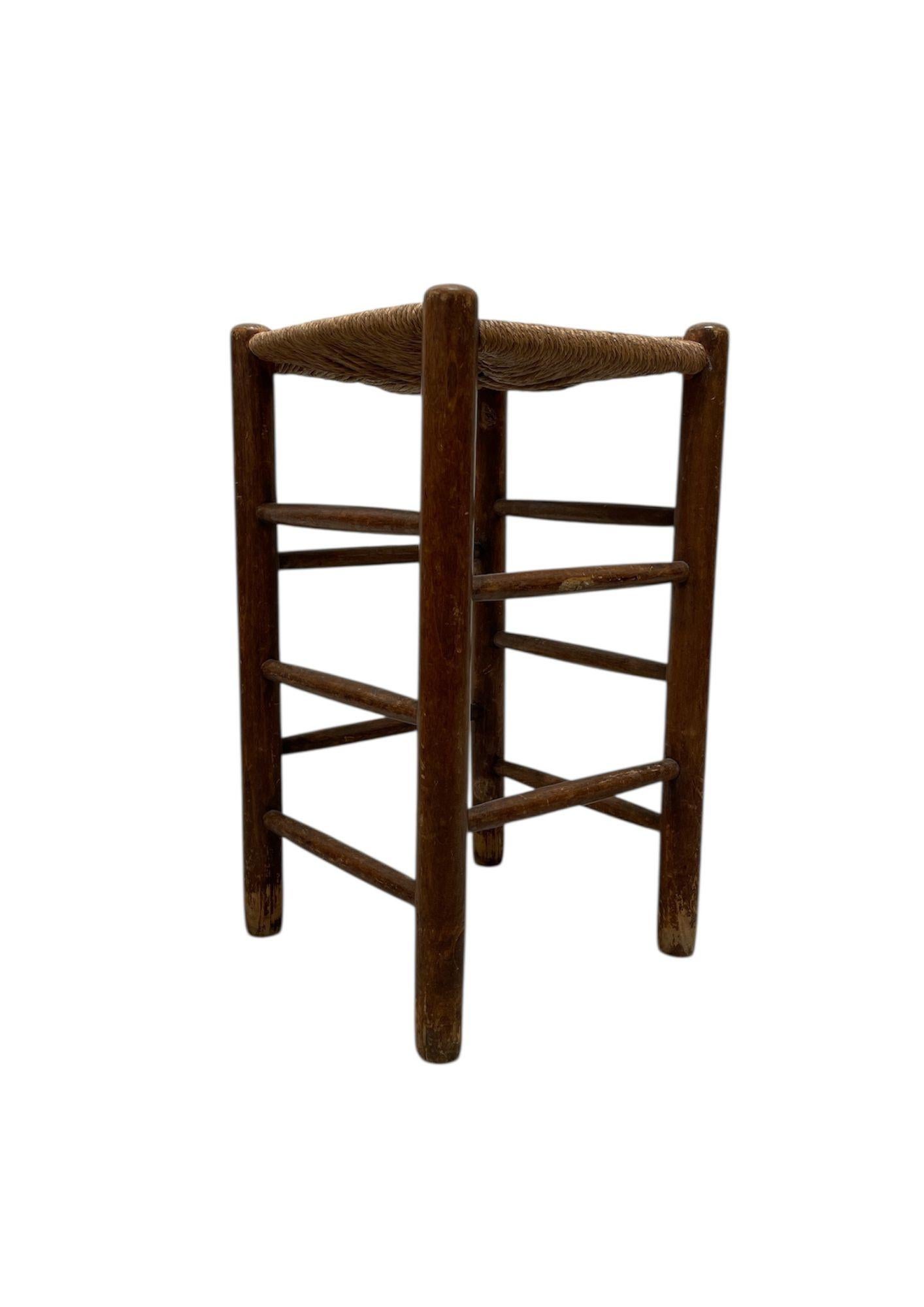 Sgabello rustico in legno patinato con seduta quadrata in paglia intrecciata con motivo a stella. La struttura, composta da montanti rotondi collegati da traverse, presenta un'usura autentica che ne testimonia l'età e il fascino originale.