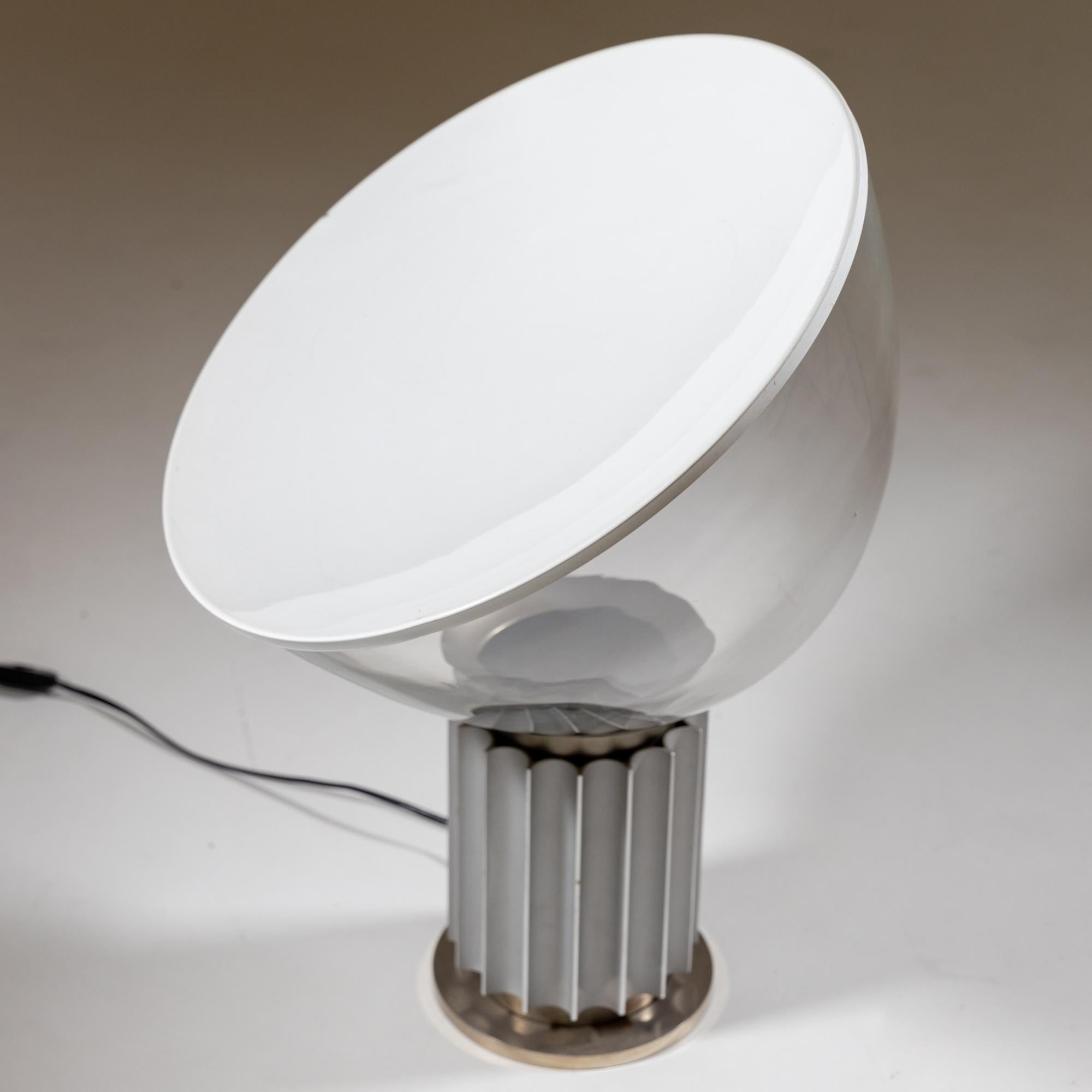 Lampe de table modèle Taccia, qui a été conçue par Achille et Pier Giacomo Castiglioni pour Flos dans les années 1960. La lampe repose sur une base ronde cannelée, sur laquelle est posé en biais le grand abat-jour en verre avec un disque concave