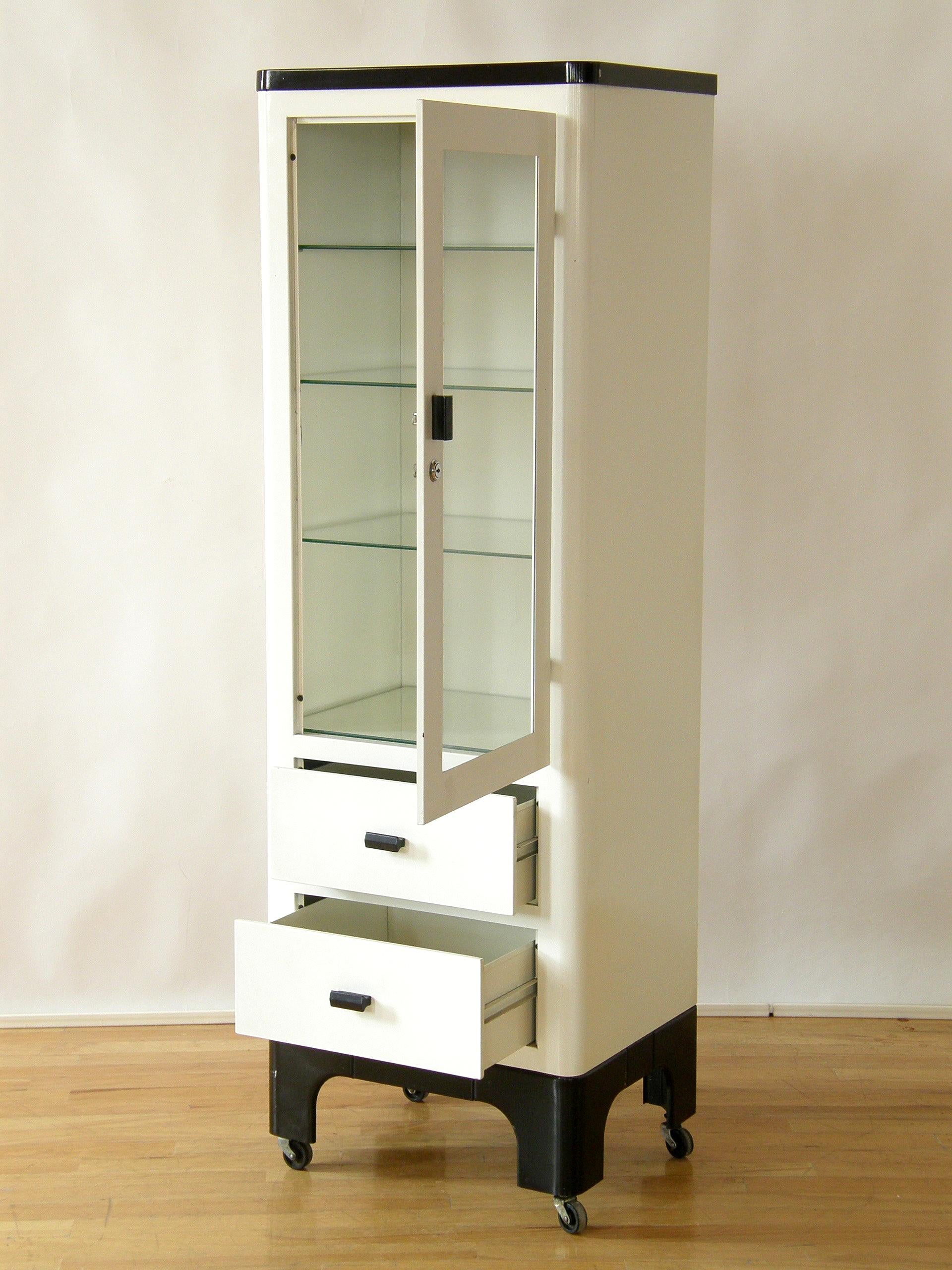 This tall, circa 1930s medical cabinet has a Machine Age, modernist style with sleek rounded corners, a white enameled steel body, and black enamel top and base. The storage includes an area with three adjustable glass shelves behind a locking,