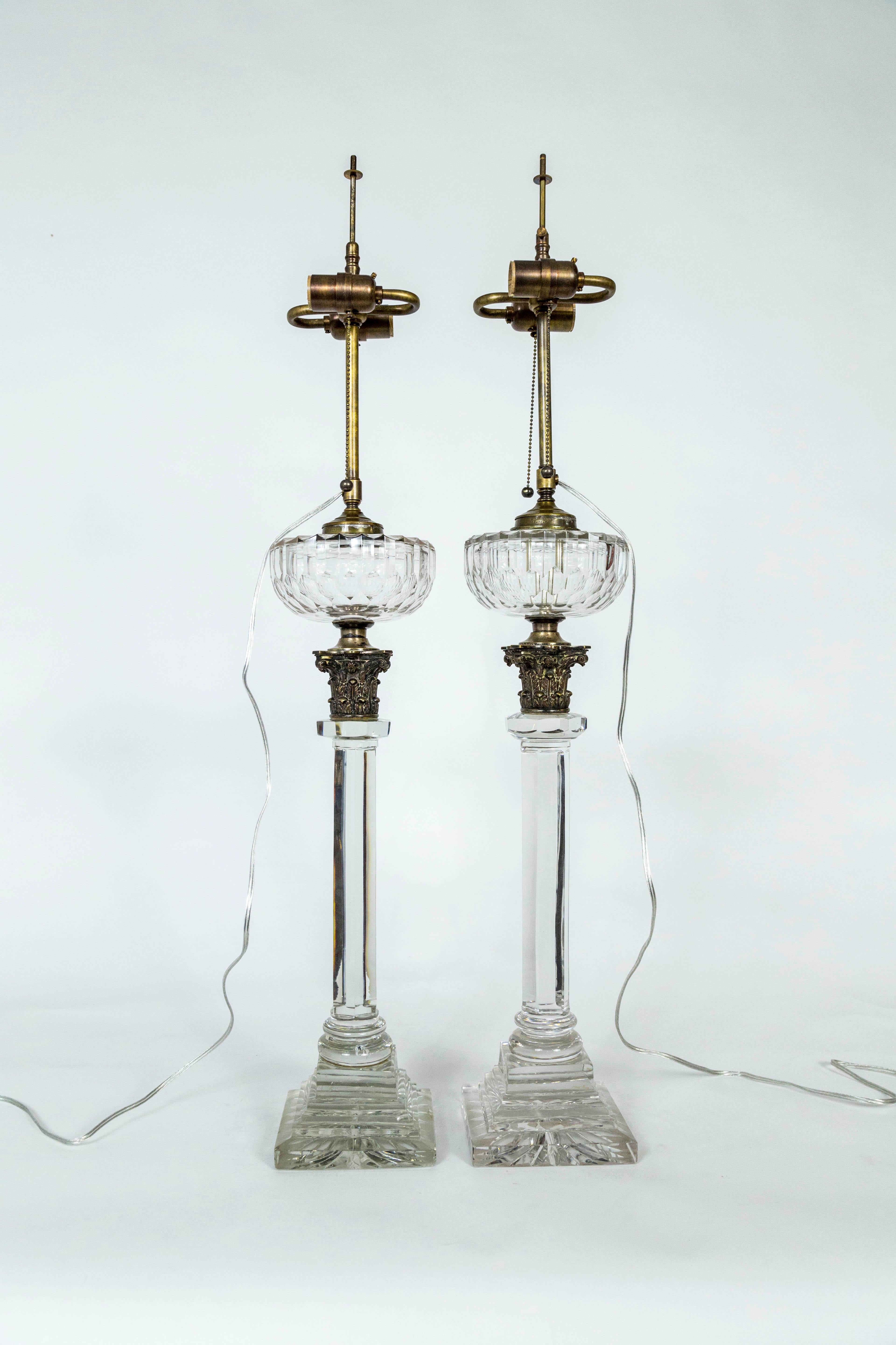 A pair of electrified former oil lamps in solid crystal columns with bronze, corinthian capitals. Cut crystal details and neoclassical elements, with brass hardware, double sockets, and pull chains; newly wired. Hinks & Sons patent, mid-19th