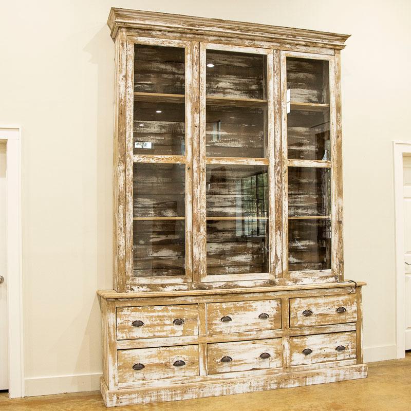 This charm of bookcase comes in the clean straight lines that accent the long glass display doors. The weathered white paint has been scraped, revealing the natural wood below adding to the French country appeal of this large cabinet. It is