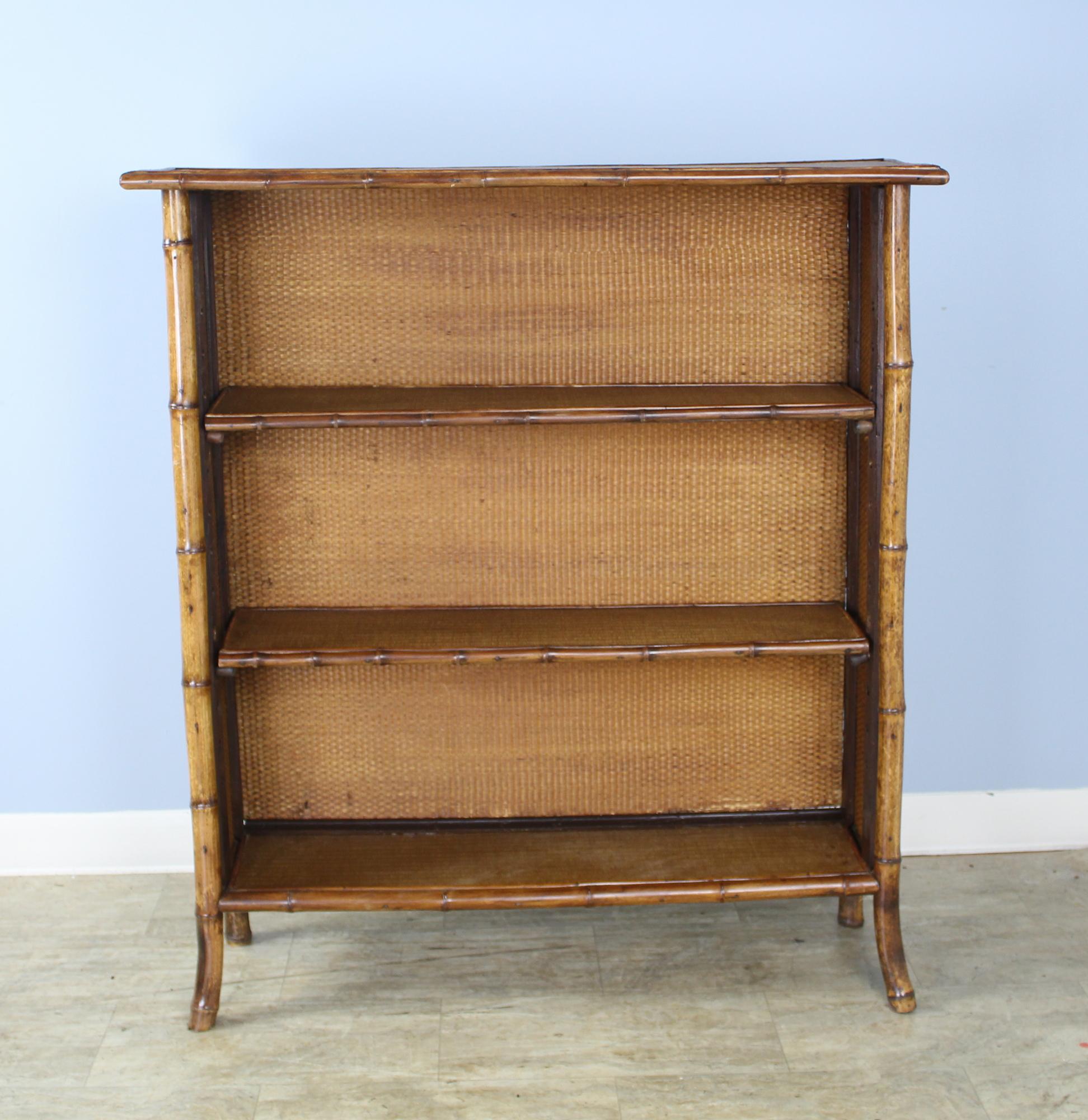 A sleek bamboo and rattan bookcase with adjustable shelves and flared front legs. The rattan is in good antique condition, with small area of wear near the bottom, shown in image #11. Pressed leather sides with floral motif are also in good