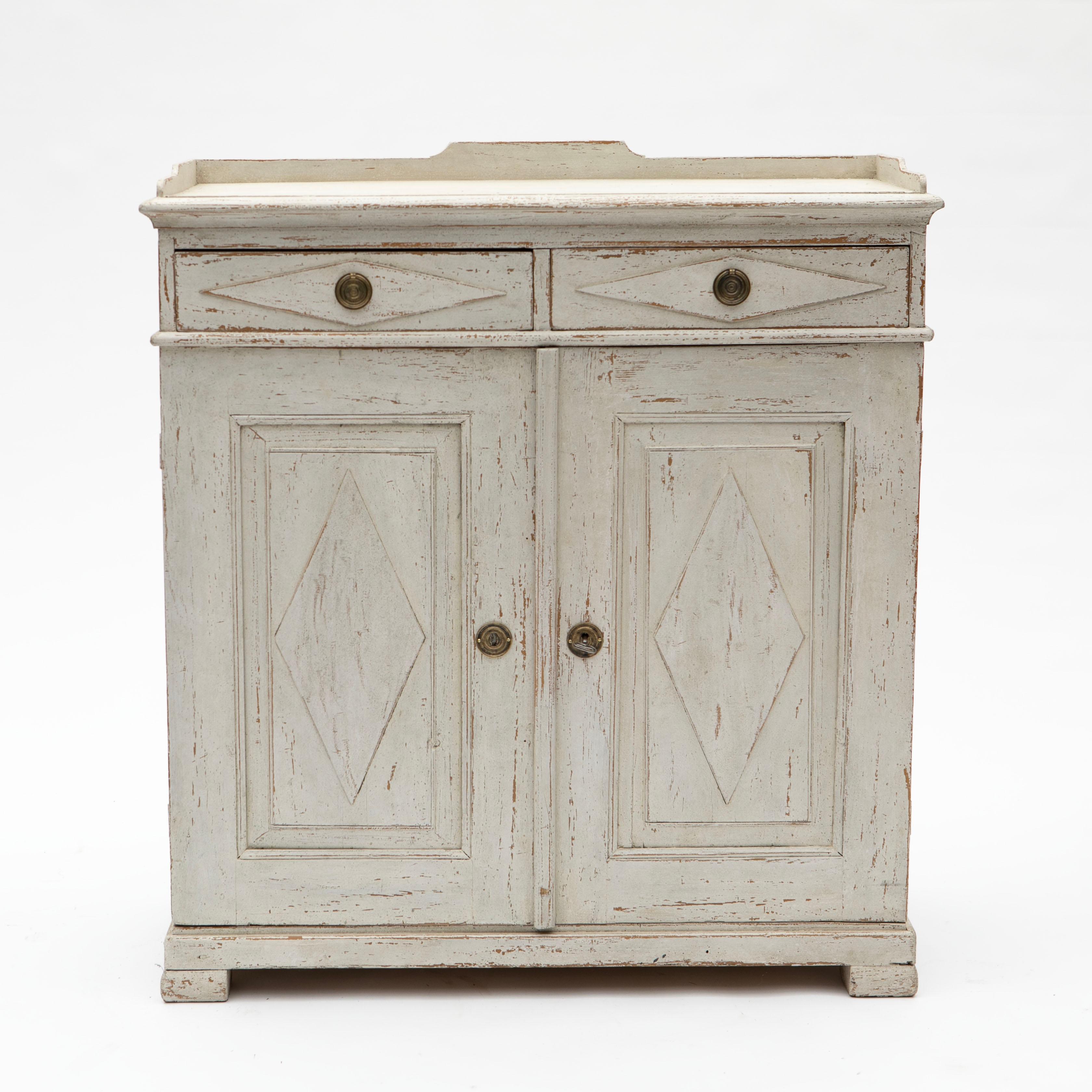 A tall antique Swedish Gustavian style sideboard or cupboard crafted in pine and later professionally painted in light grey with authentic distress and patina.
wo top drawers, each with carved diamond mouldings and brass fittings.
Below the drawers