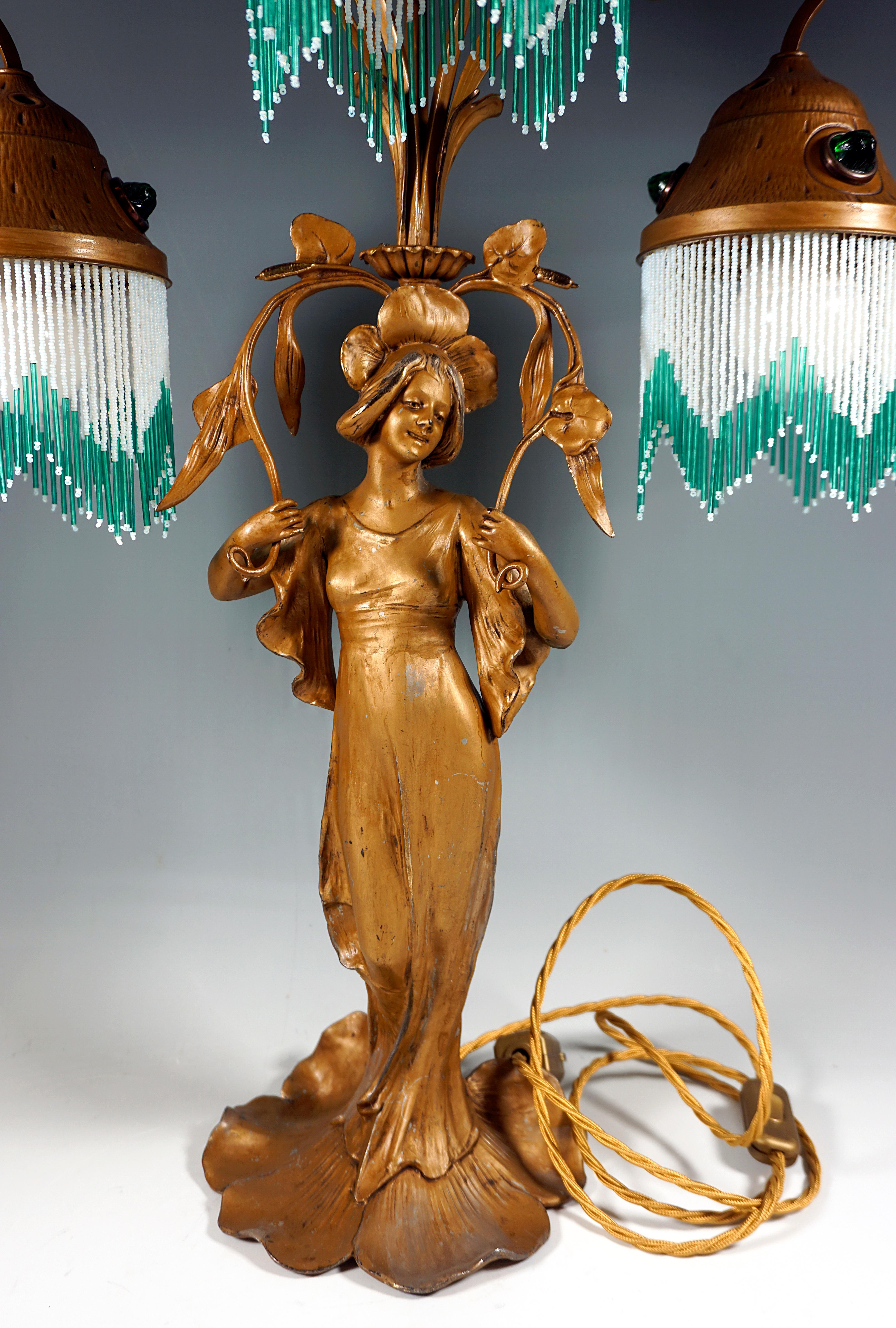 Tall table lamp with a young woman decorated with flowers, 'Flora' - goddess of blooming, as a base, stylized branch-like and curved struts originating behind her head with three round shades with green gemstones and strings of beads decoration,