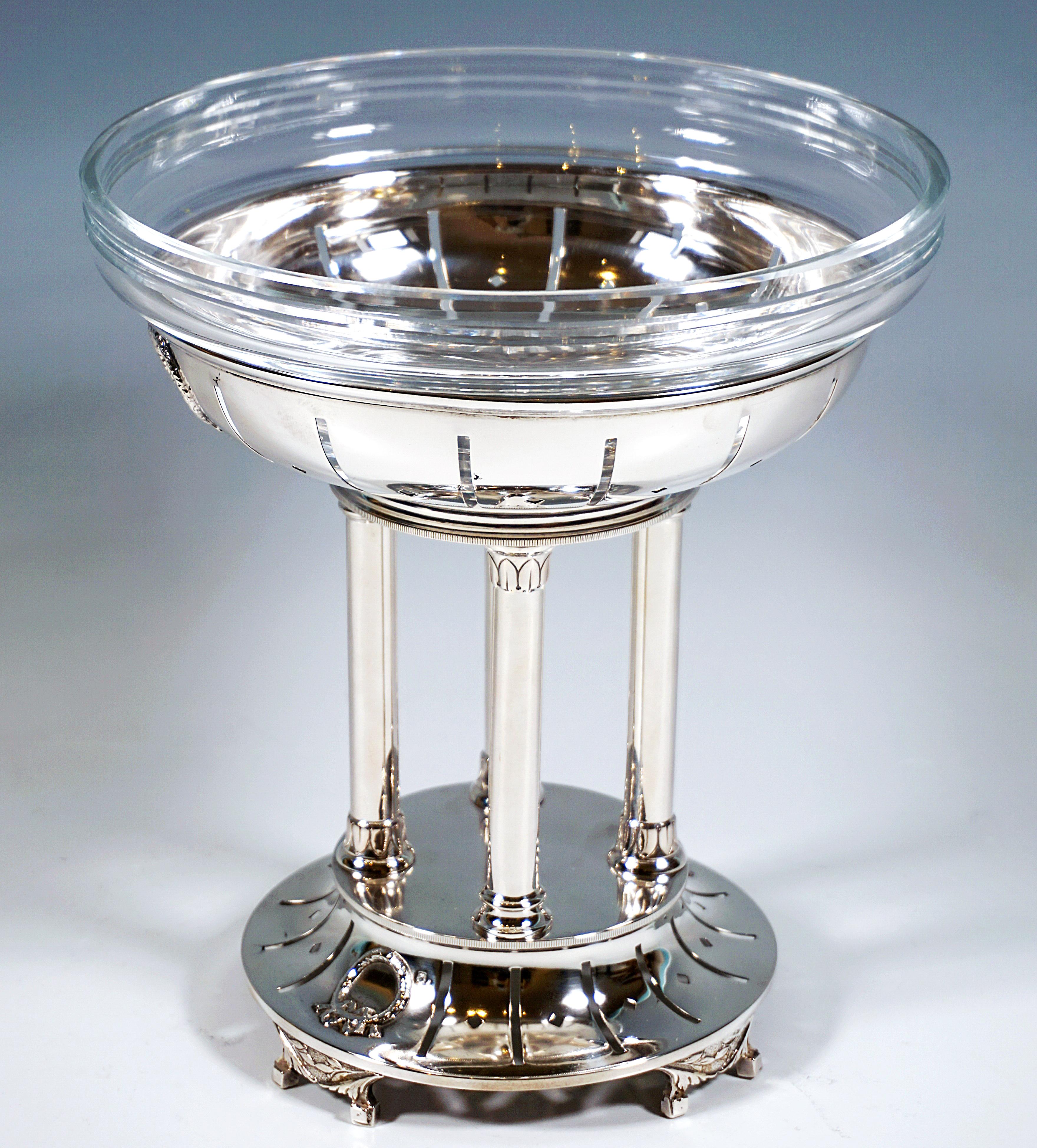 Elegante centro de mesa de plata sobre una base alta, redonda, estriada y escalonada sobre cuatro pies divididos con decoración floral, con perforaciones radiales alargadas y alternadas en forma de pequeños rombos en las estrías, dos coronas en