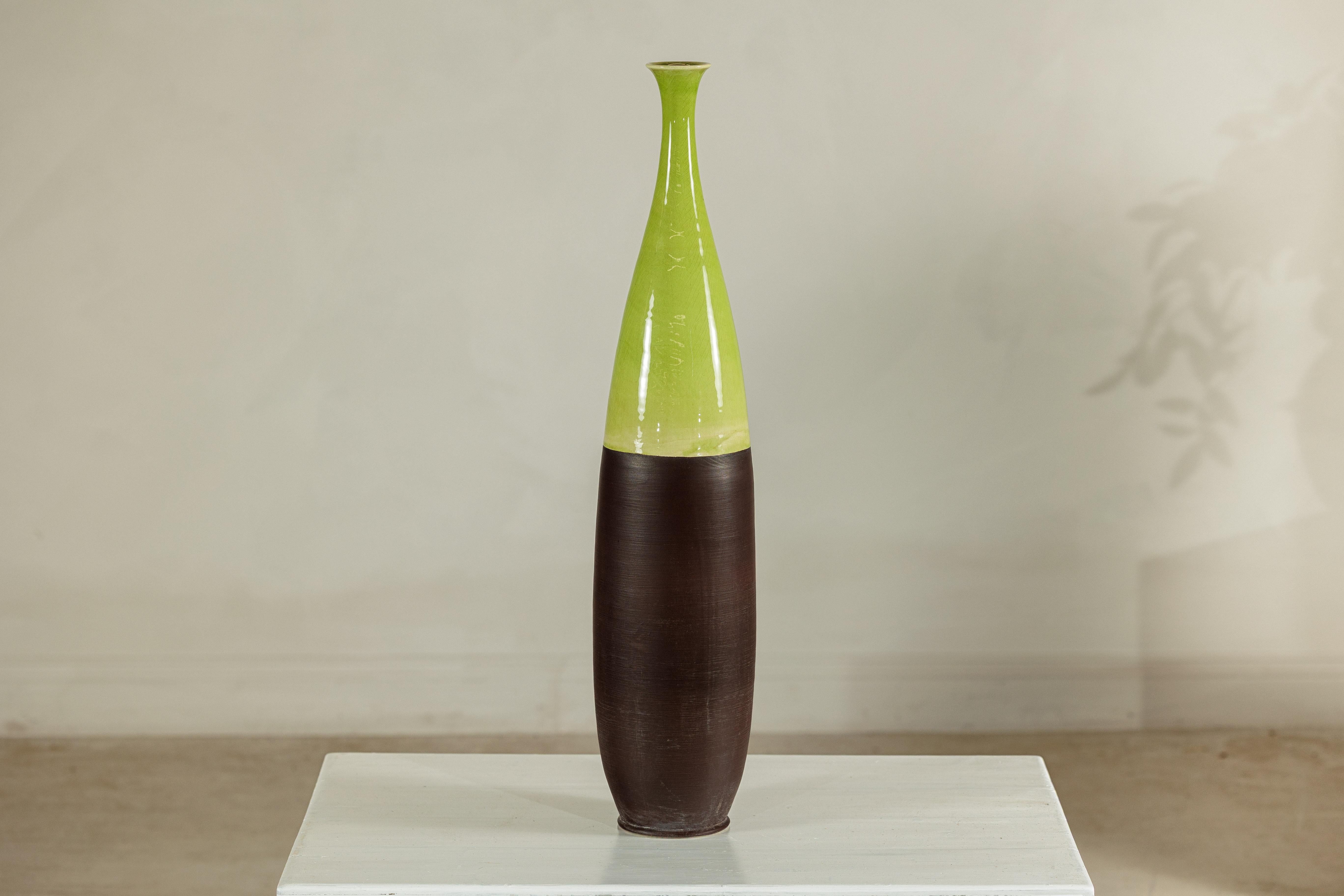 This tall artisan ceramic vase captivates with its elegant, elongated form and refined two-tone glaze. The glossy upper section, finished in a vivid yellow-green, contrasts beautifully with the deep matte brown lower half, creating a bold visual